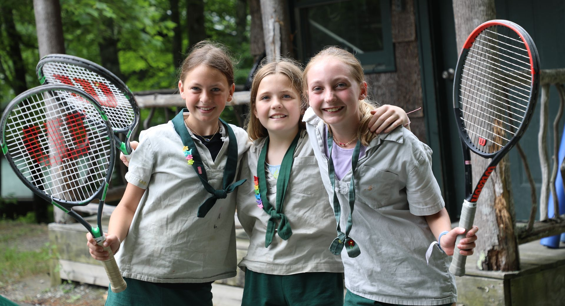 Three children in scout uniforms with green neckerchiefs smile while holding tennis rackets outdoors.
