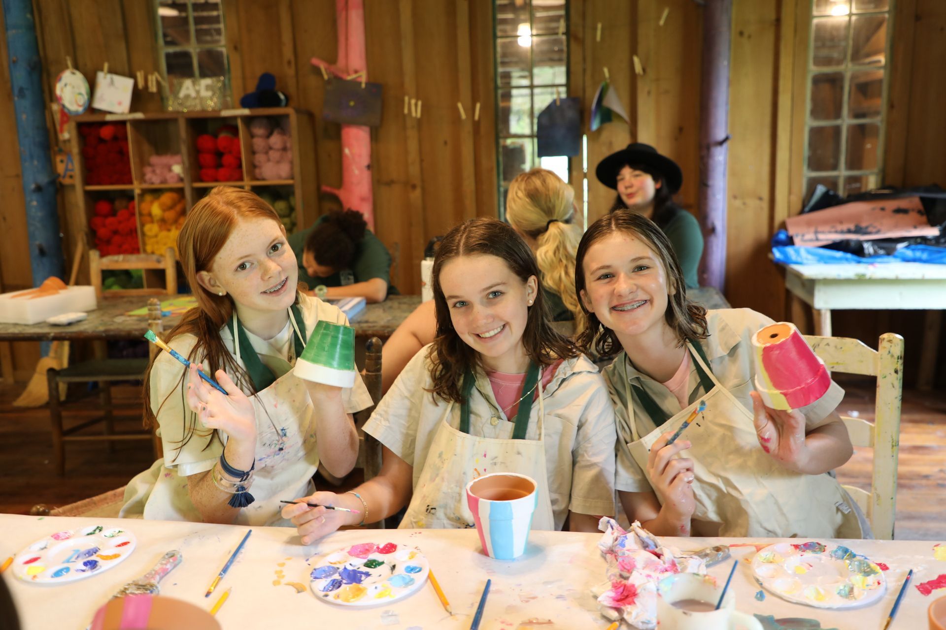 Three people wearing aprons sit at a table, smiling while painting small clay pots in a craft room filled with art supplies.