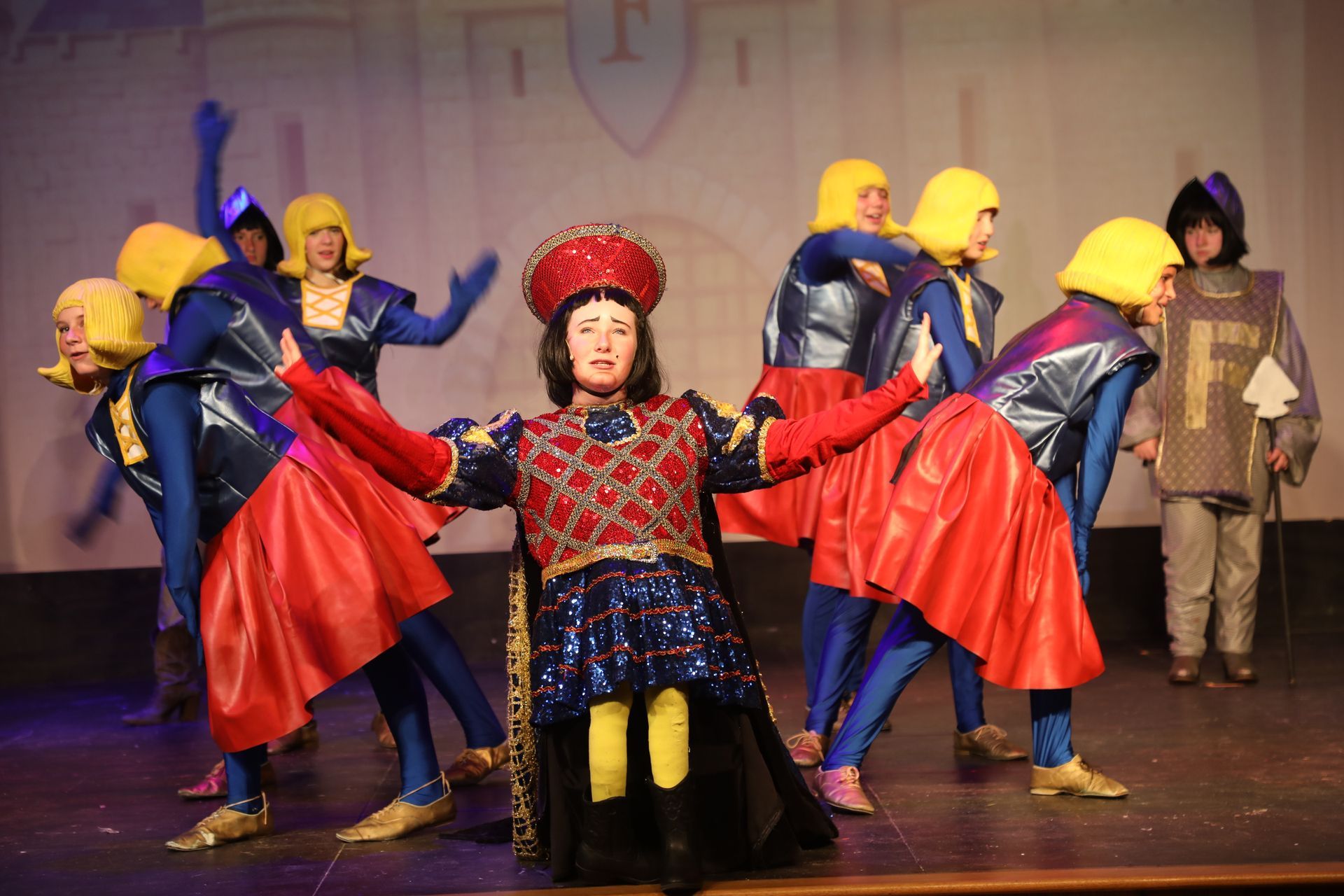 A theater performance showing Lord Farquaad kneeling center stage surrounded by performers in royal blue and red costumes.