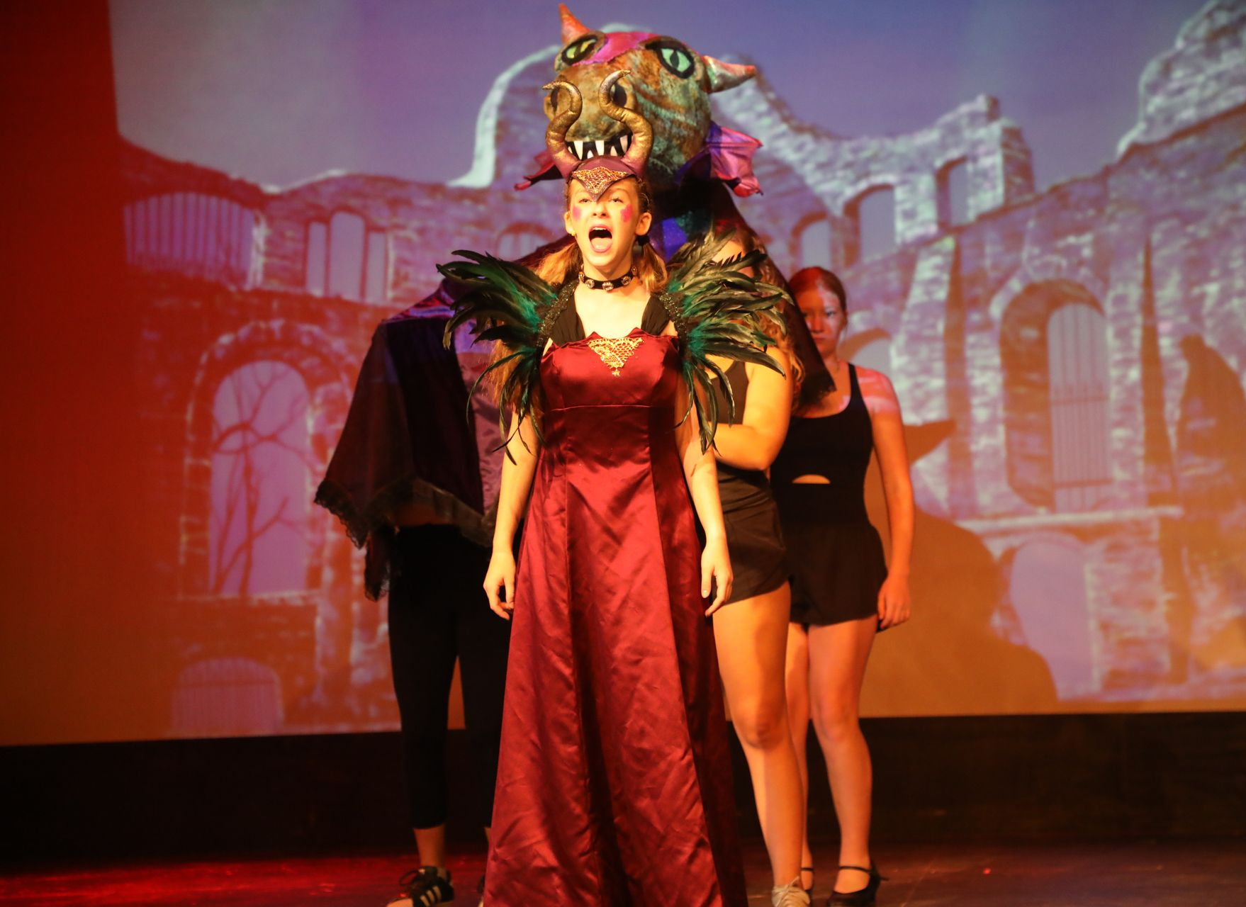 Actors on a stage perform in front of a projection of ruins; the central figure wears a red dress and an animal mask.