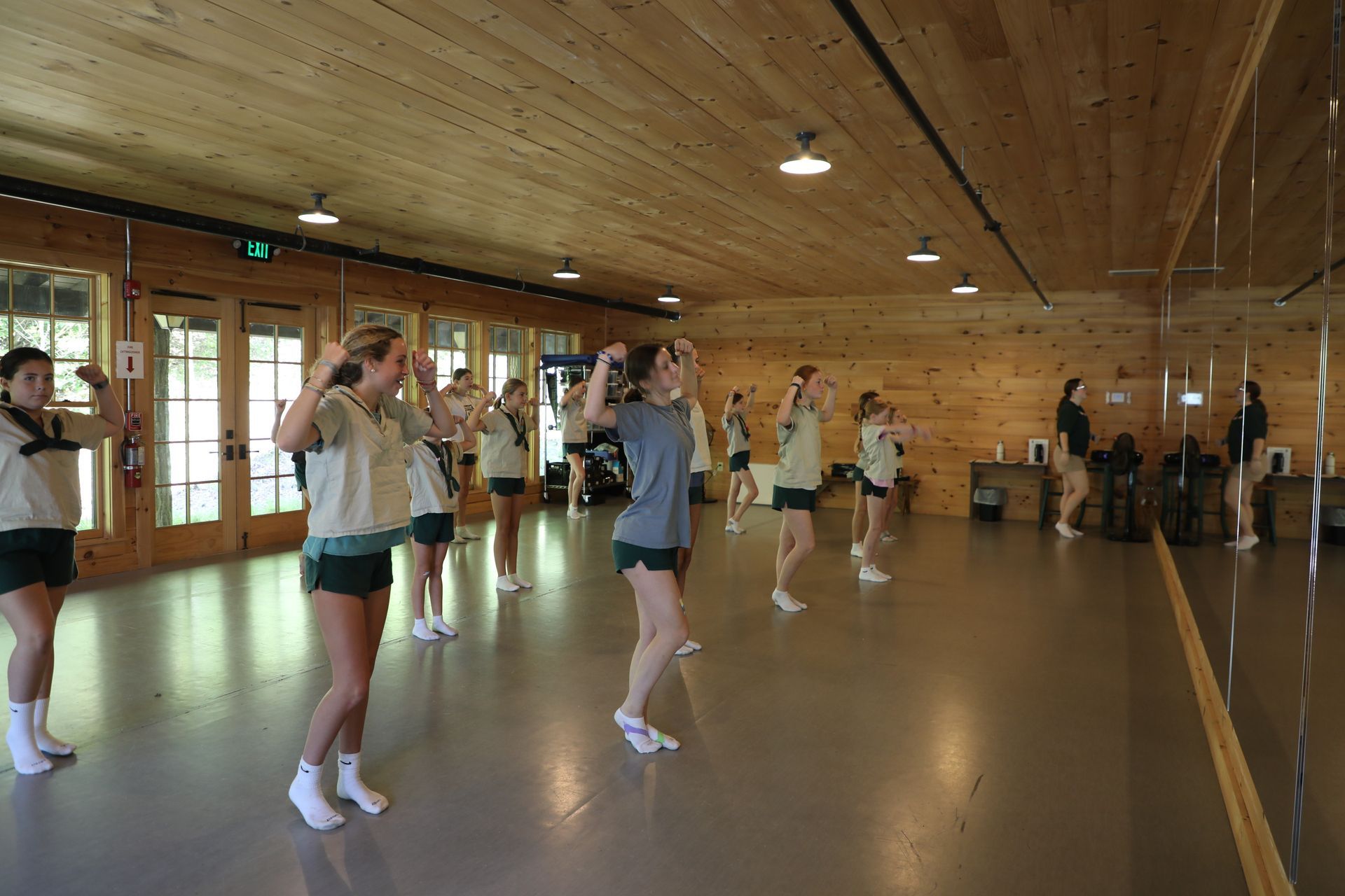 A group of people wearing athletic clothes stand in a line in a wood-paneled dance studio, posing with arms flexed.