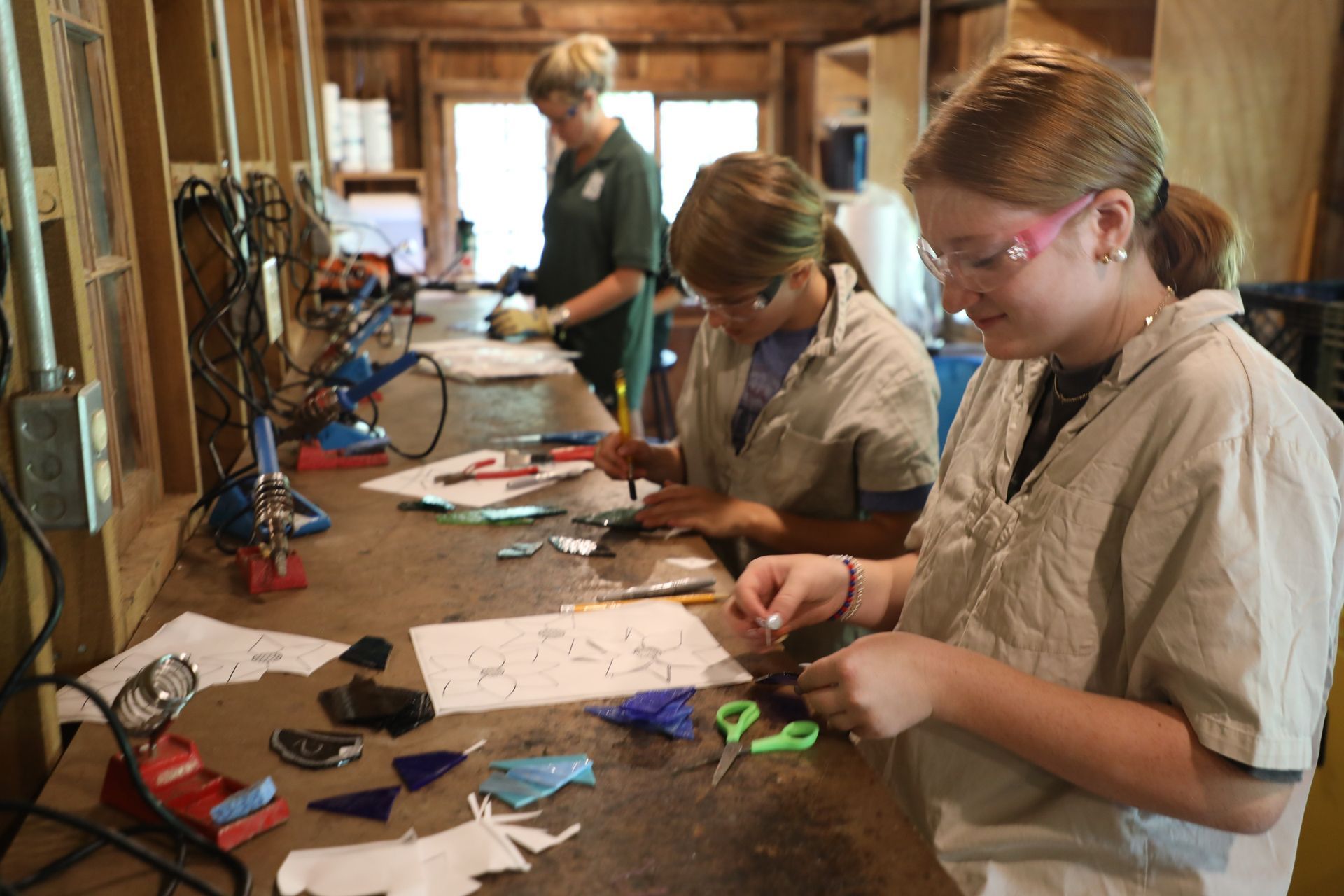 Three people in a workshop focus on assembling stained glass pieces at a long wooden table.