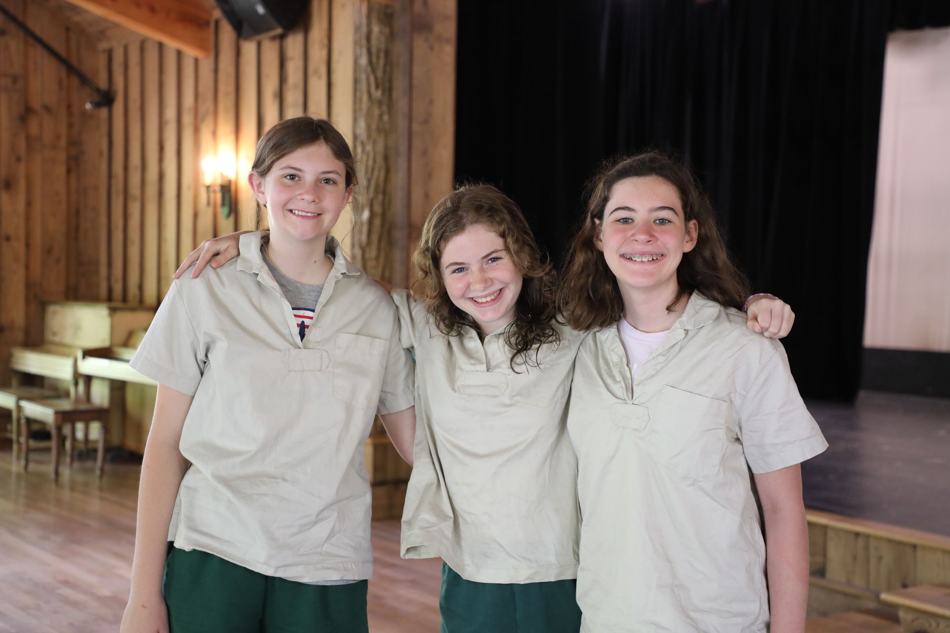 Three people in light-colored uniform shirts and green bottoms stand smiling with arms around each other in a hall.