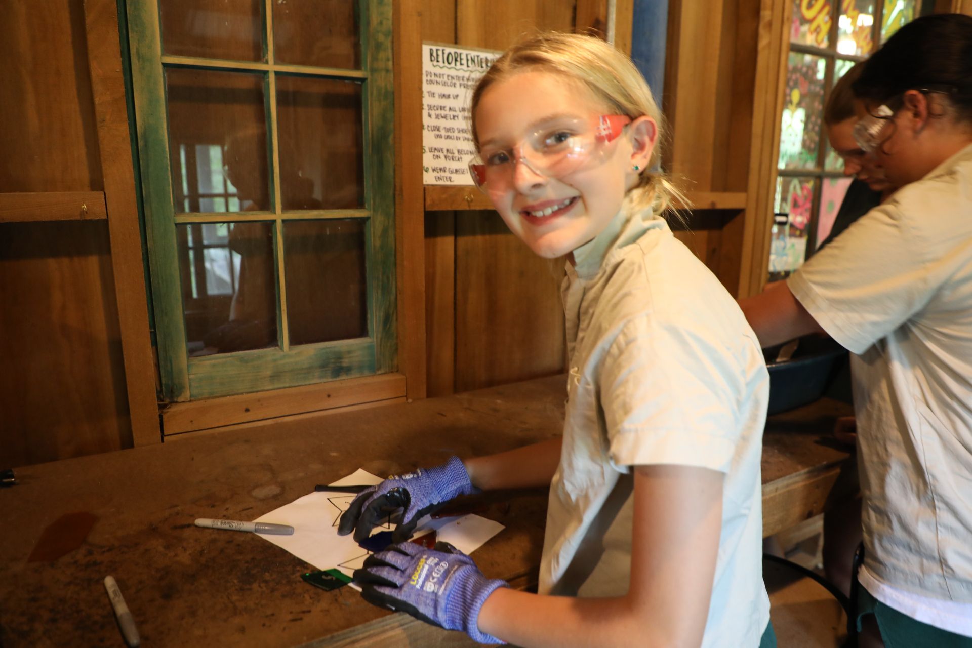 A person wearing safety glasses and work gloves smiles while working with materials on a wooden table in a workshop.