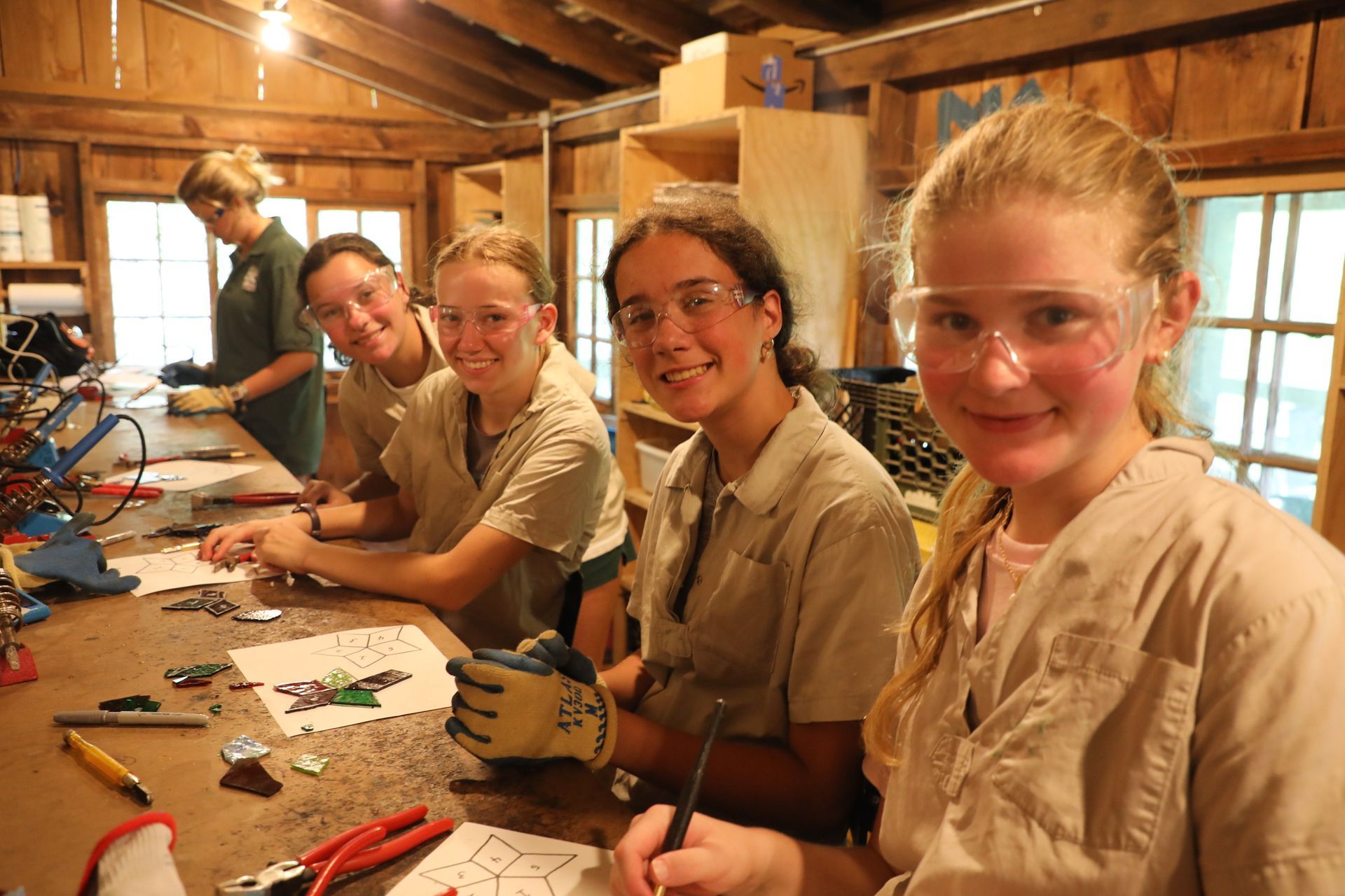 Four people wearing safety goggles and work shirts crafting at a wooden workbench in a workshop.