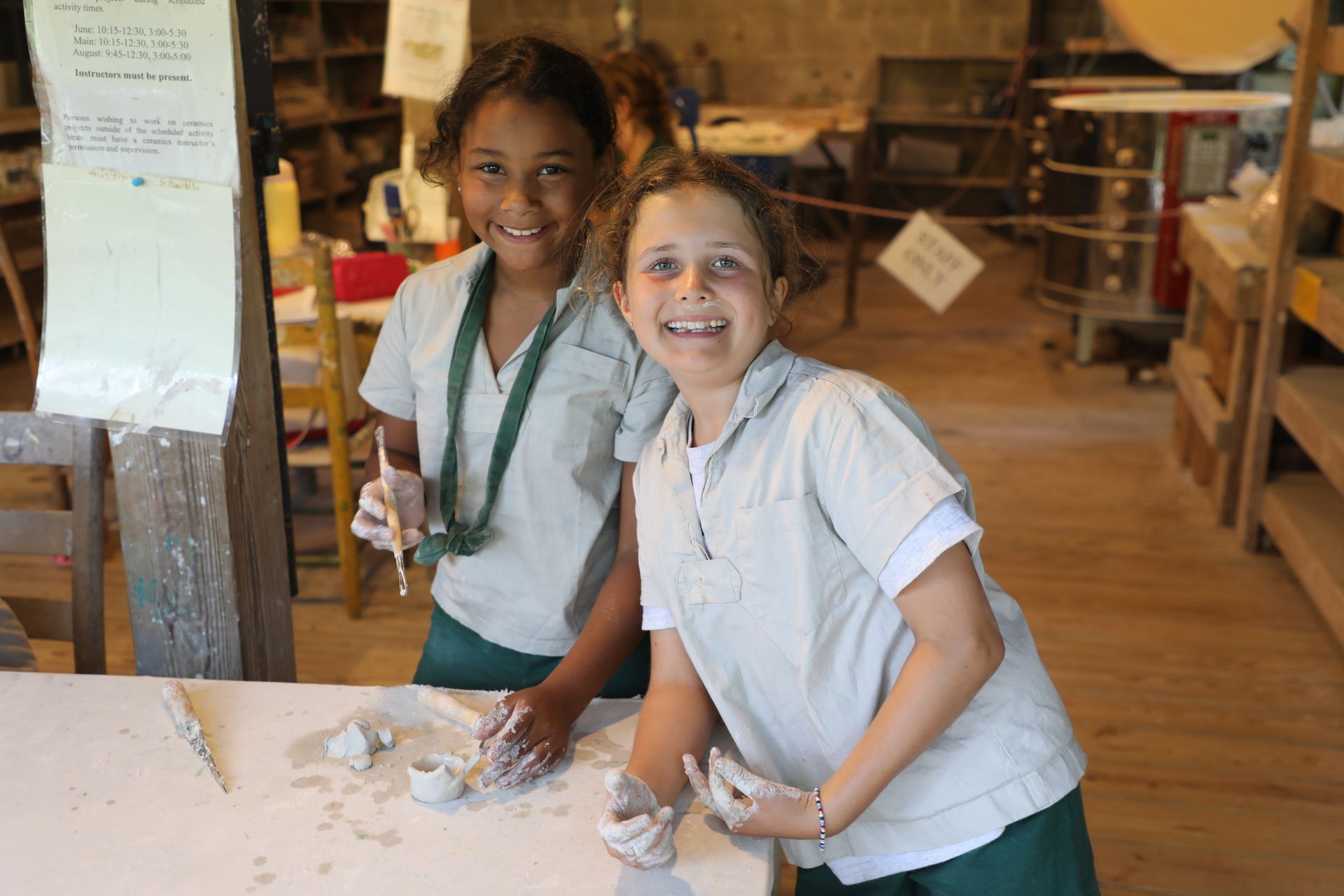 Two children in uniforms smiling while working with clay at a table in a pottery workshop.
