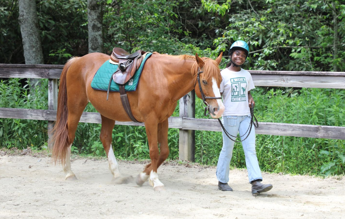A person wearing a helmet leads a saddled chestnut horse along a dirt path beside a wooden fence in a wooded area.