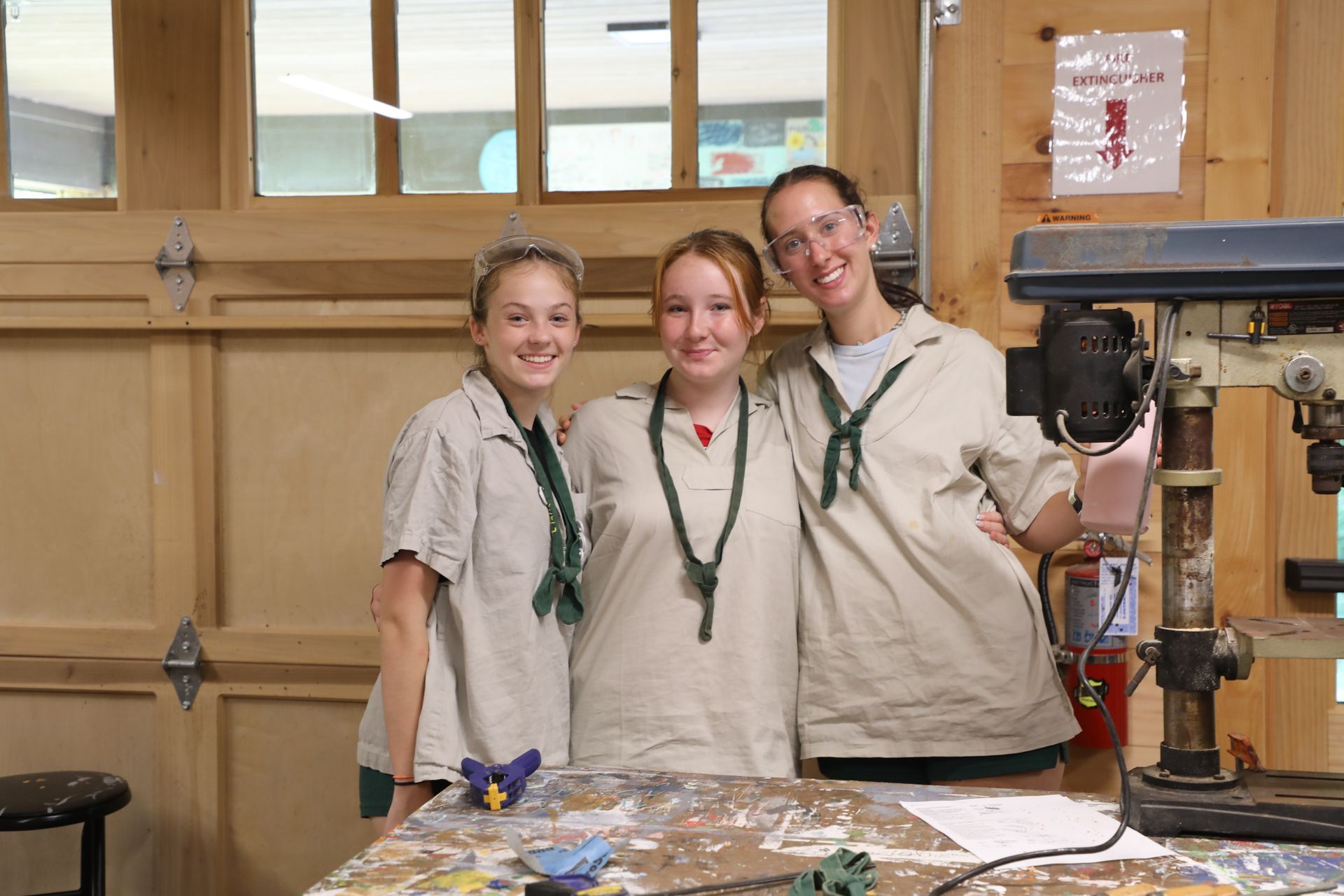 Three smiling people in beige shirts and green scarves stand together in a workshop with a drill press in the foreground.