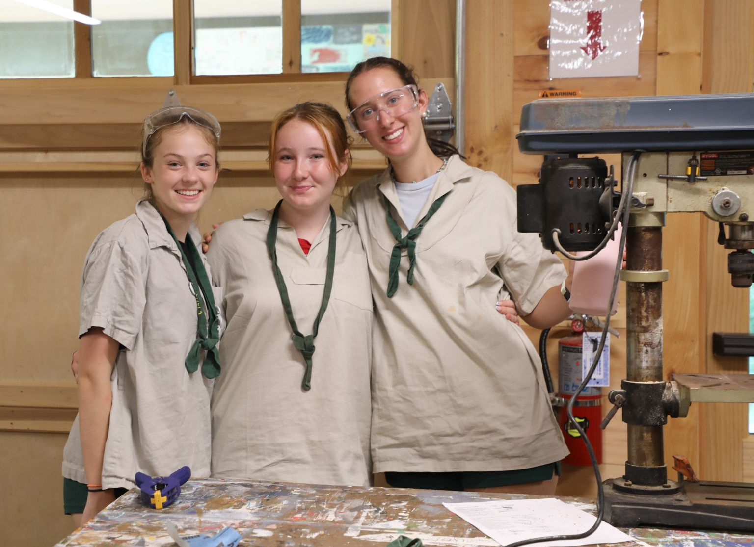 Three people in matching tan shirts and green neckerchiefs pose together in a workshop with a drill press nearby.