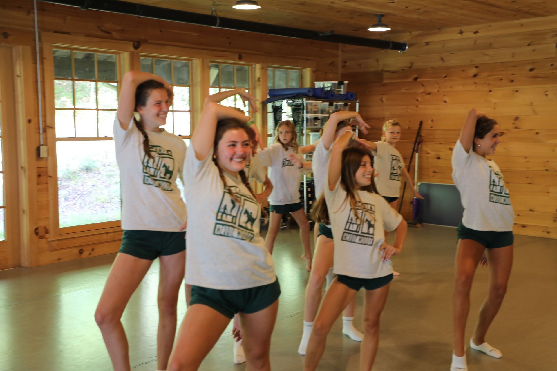 A group of dancers in white t-shirts and green shorts performing a choreographed routine in a wood-paneled room.