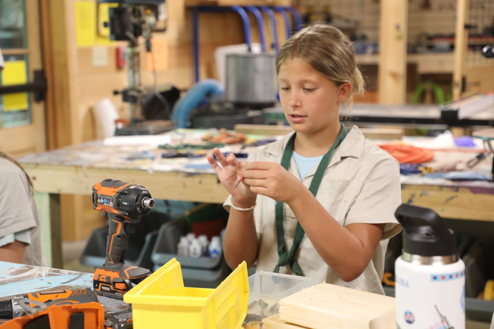 A student in a workshop holding a small screw, with tools and wooden materials on the table in front of them.