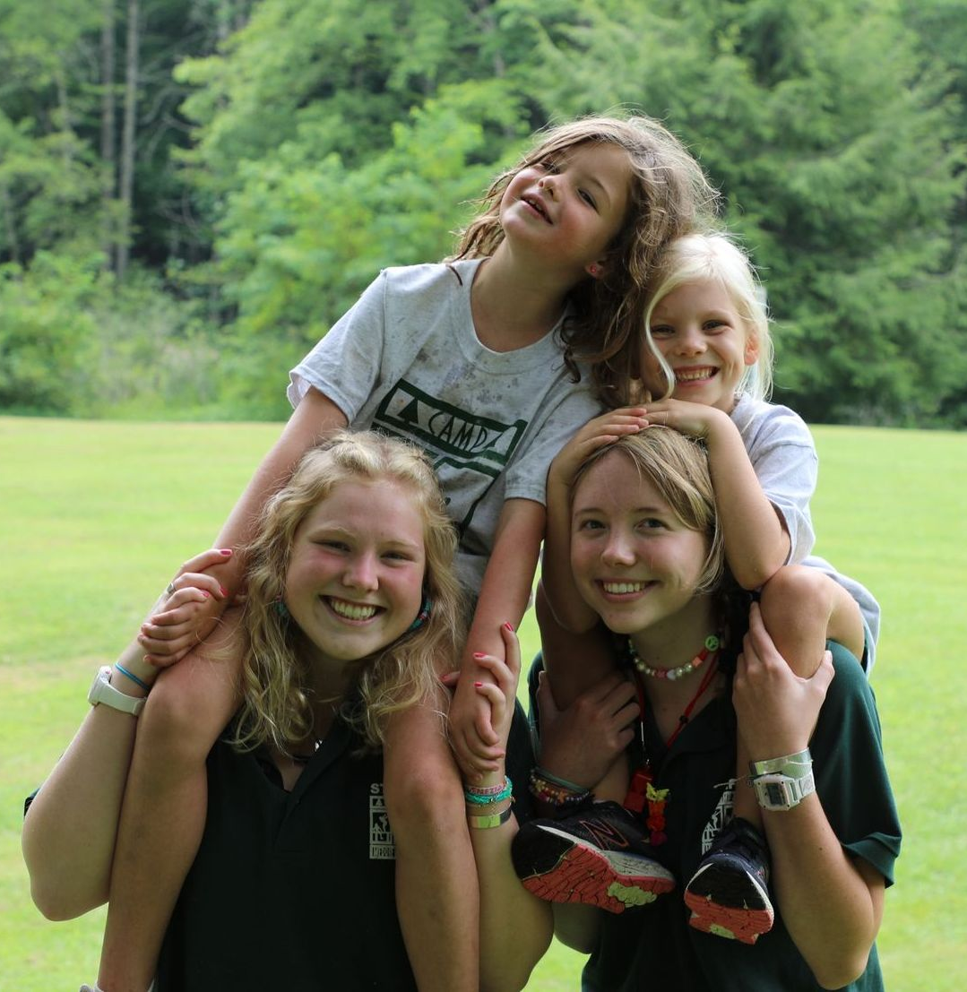 Two people carry two smiling children on their shoulders against a backdrop of green grass and trees.