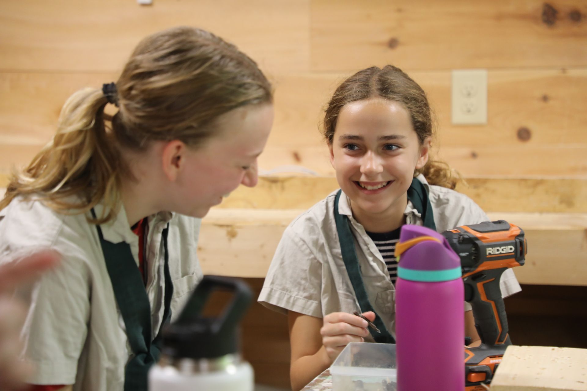 Two people in a workshop setting look at each other while smiling, with a drill and water bottle on the table.