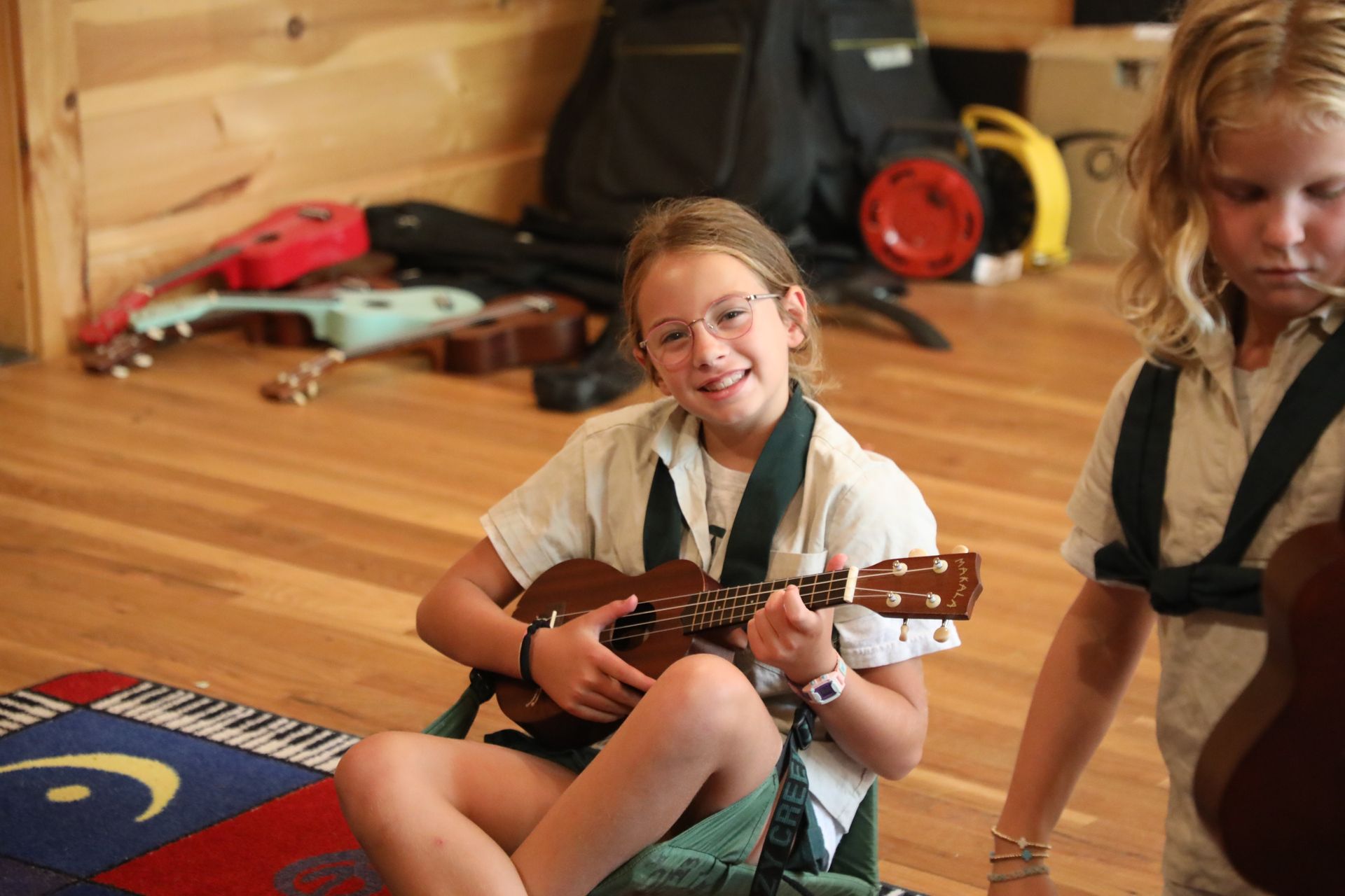A smiling child in a light shirt sits on a rug playing a small stringed instrument, with another child partially in view.