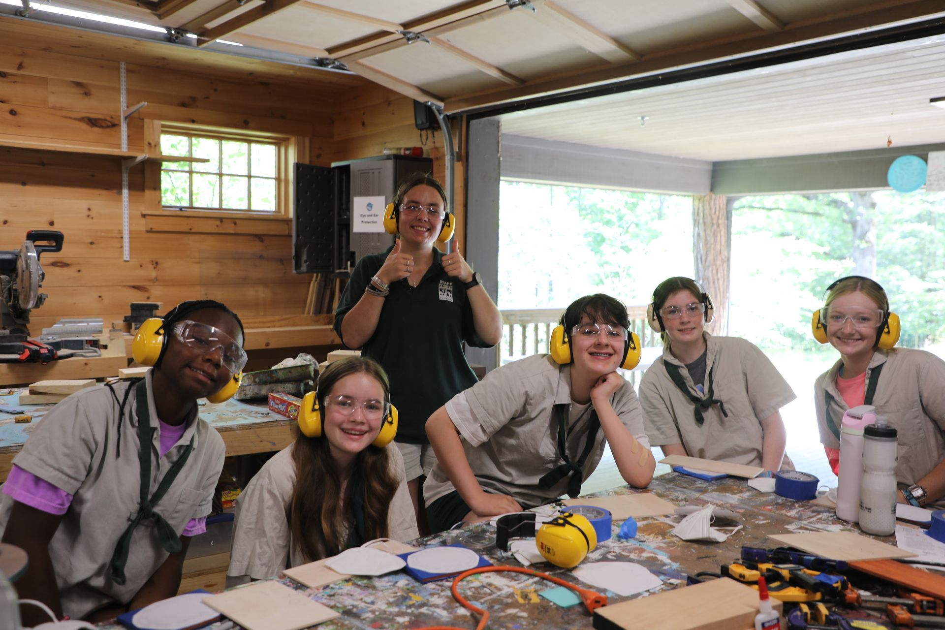 Five people wearing ear protection pose in a workshop, some smiling and others giving thumbs-up gestures.