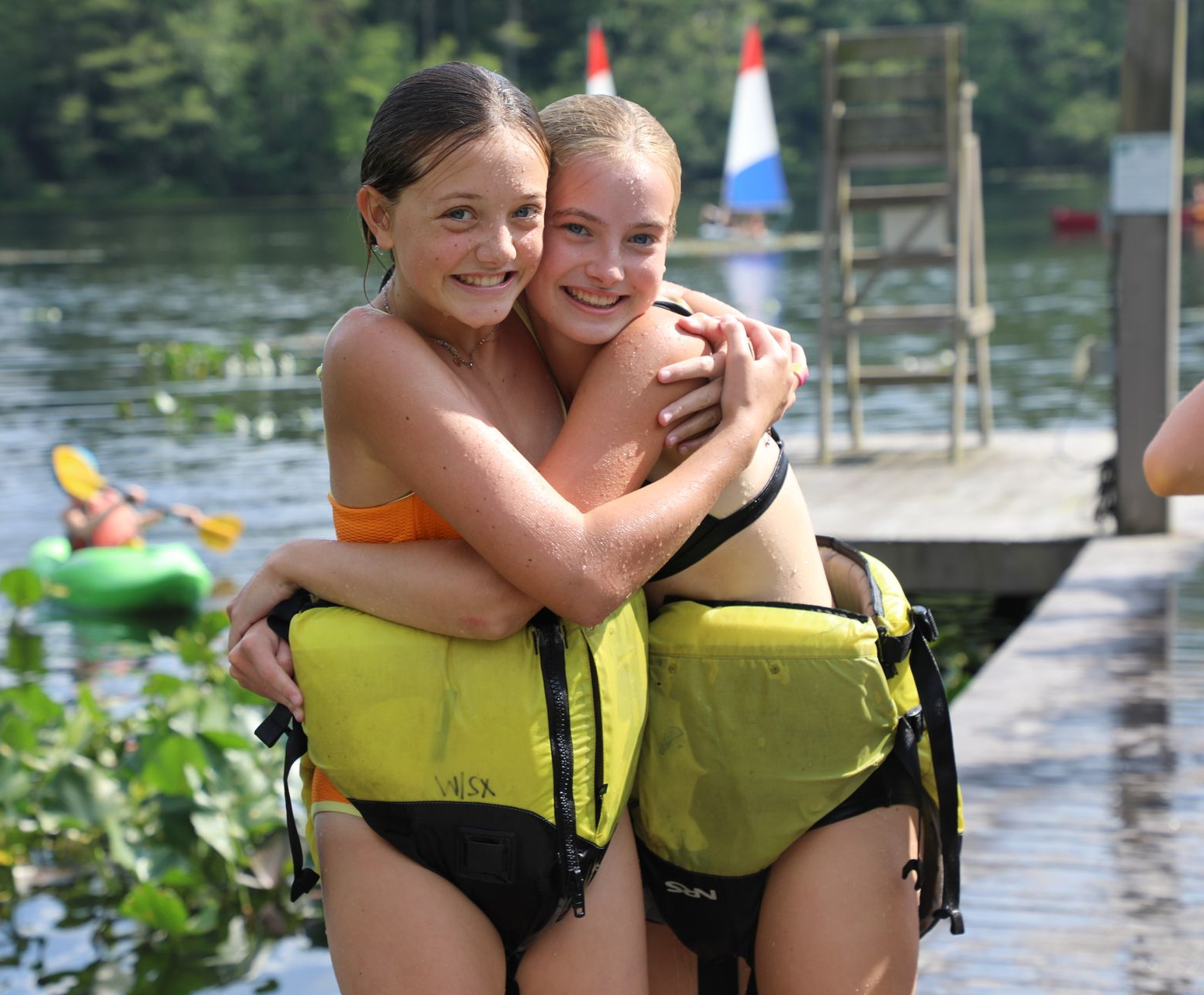 Two smiling friends wearing life vests embrace on a wooden dock by a lake, with a person kayaking in the background.