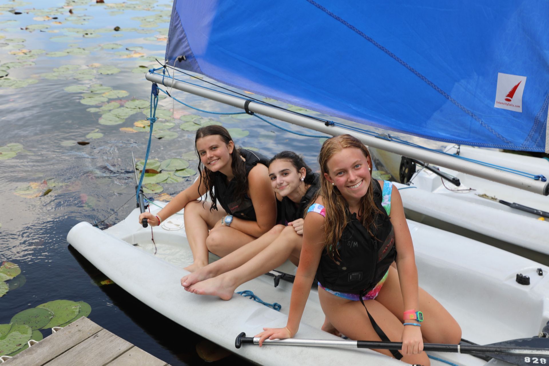 Three people in life jackets sitting in a small white sailboat with a blue sail, moored at a dock by lily pads.