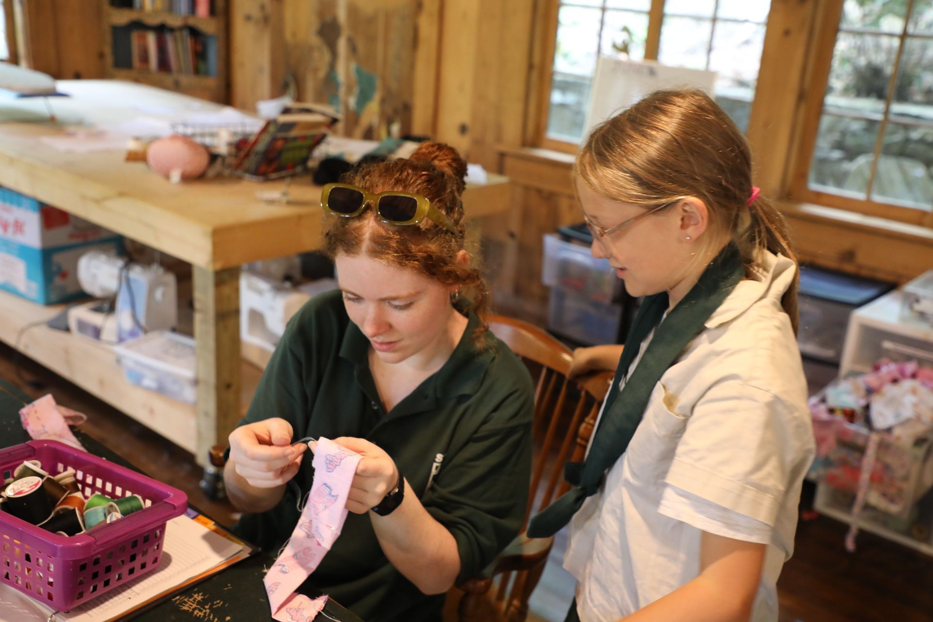 Two people in a craft room work together on a sewing project, one focused on stitching fabric while the other watches.