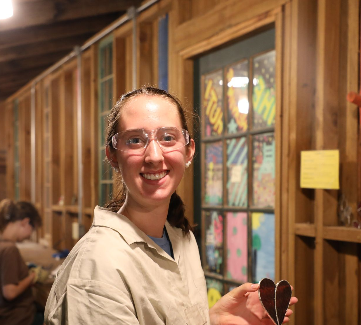A person wearing protective glasses smiles while holding a small, dark heart-shaped object in a wood-paneled room.