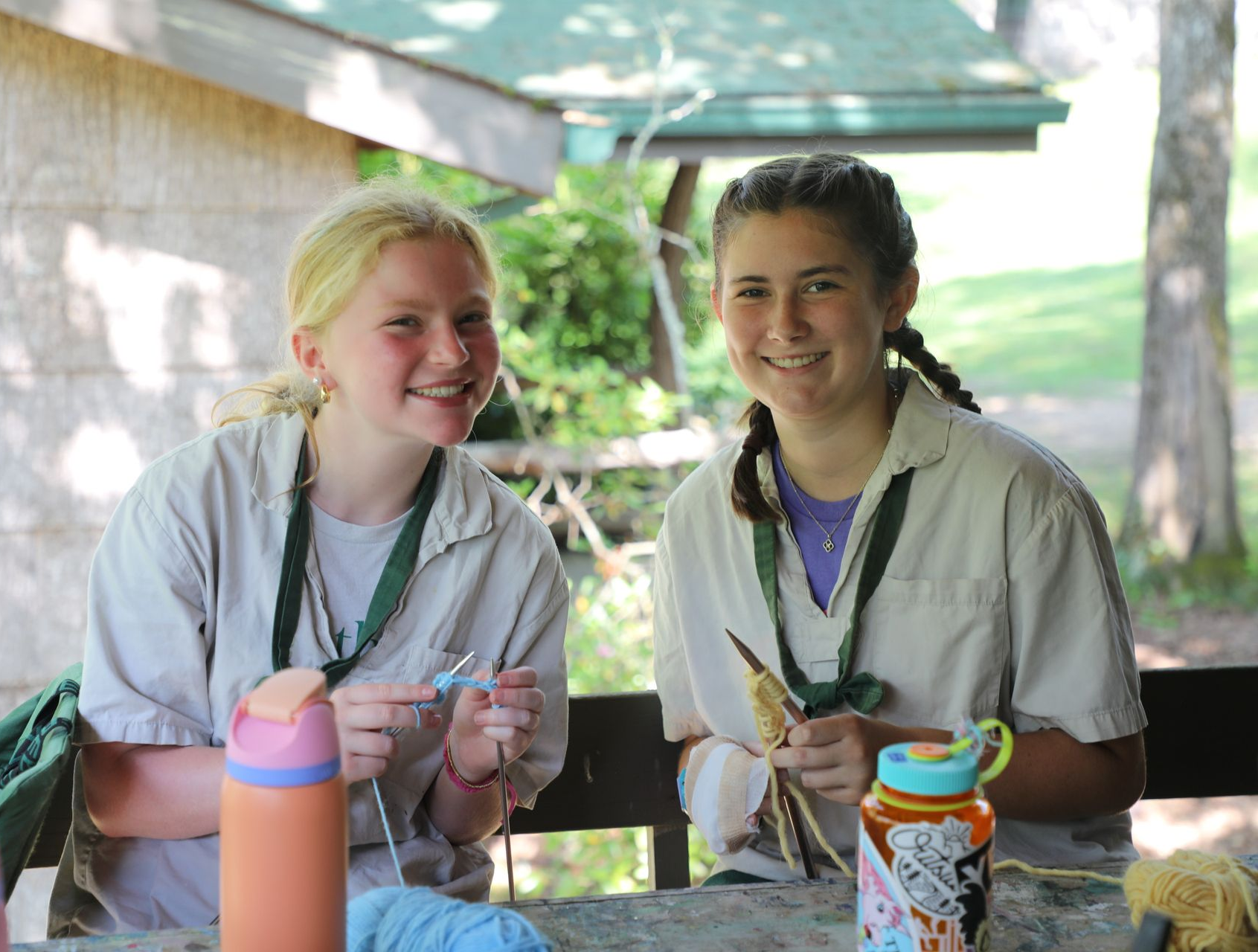 Two people in tan shirts with green neckerchiefs sit at an outdoor table, smiling while knitting with yarn.