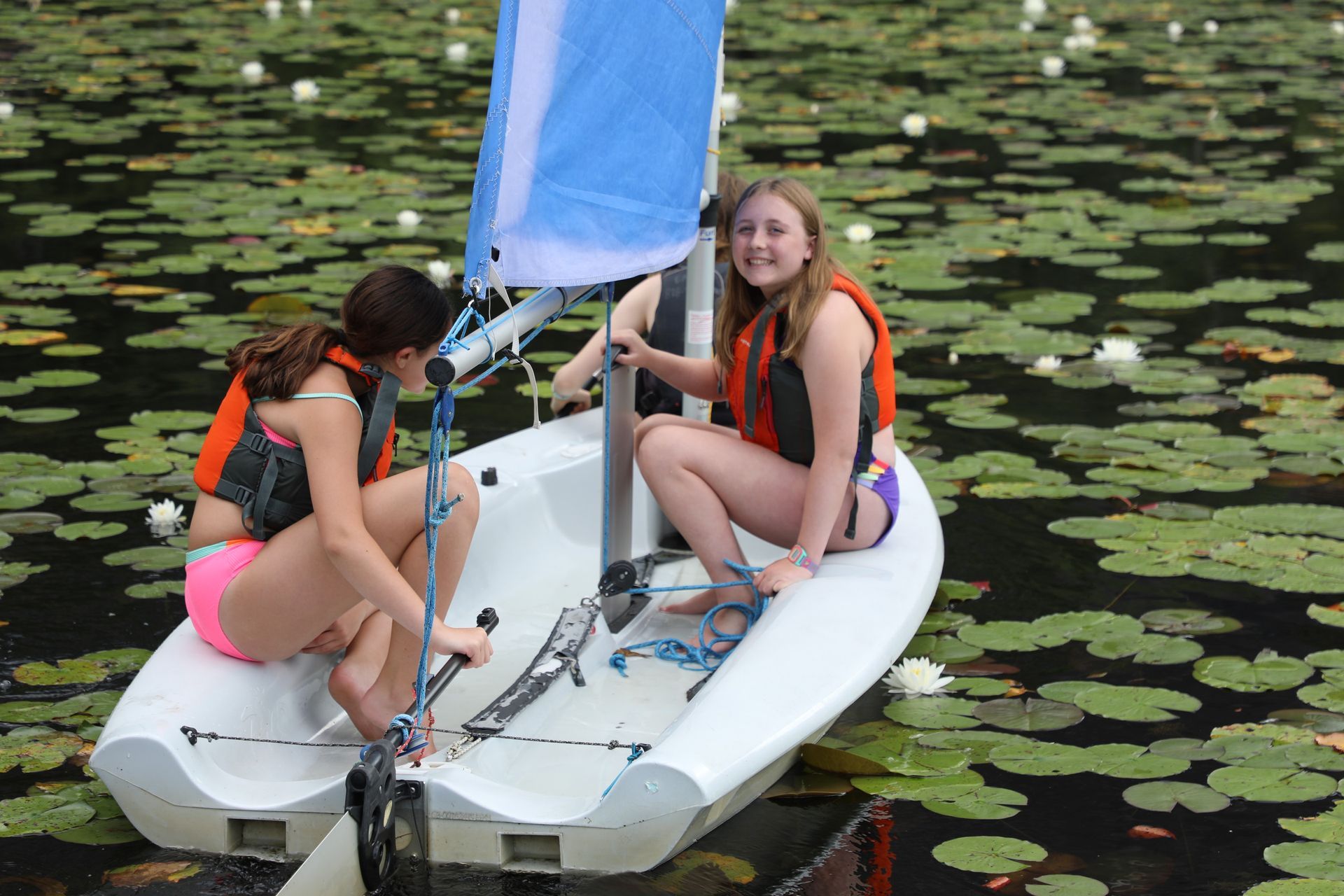 Two people wearing life vests sit in a small sailboat on a pond covered in water lilies.