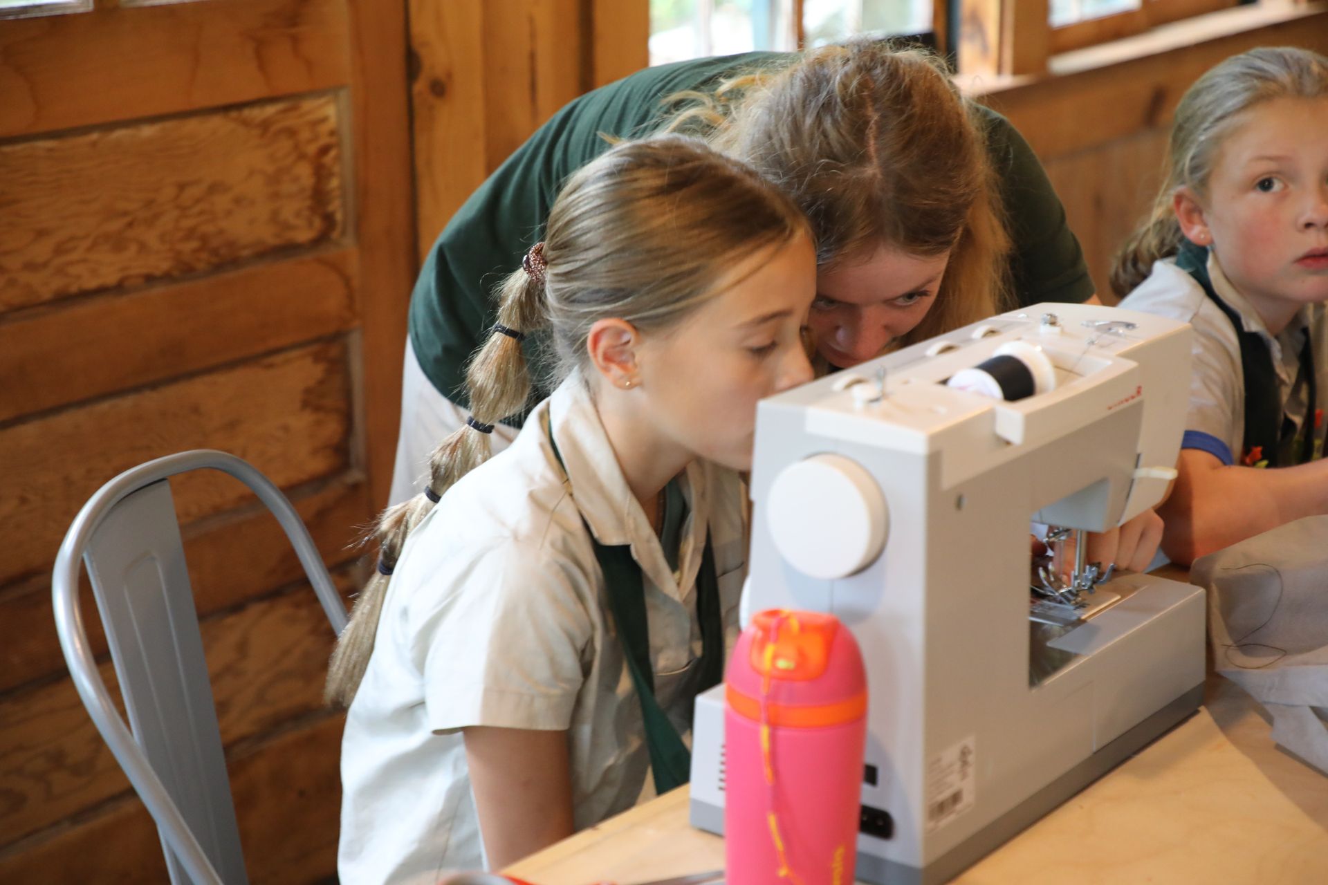 Three people in a log-walled room focusing on a sewing machine as it stitches fabric.
