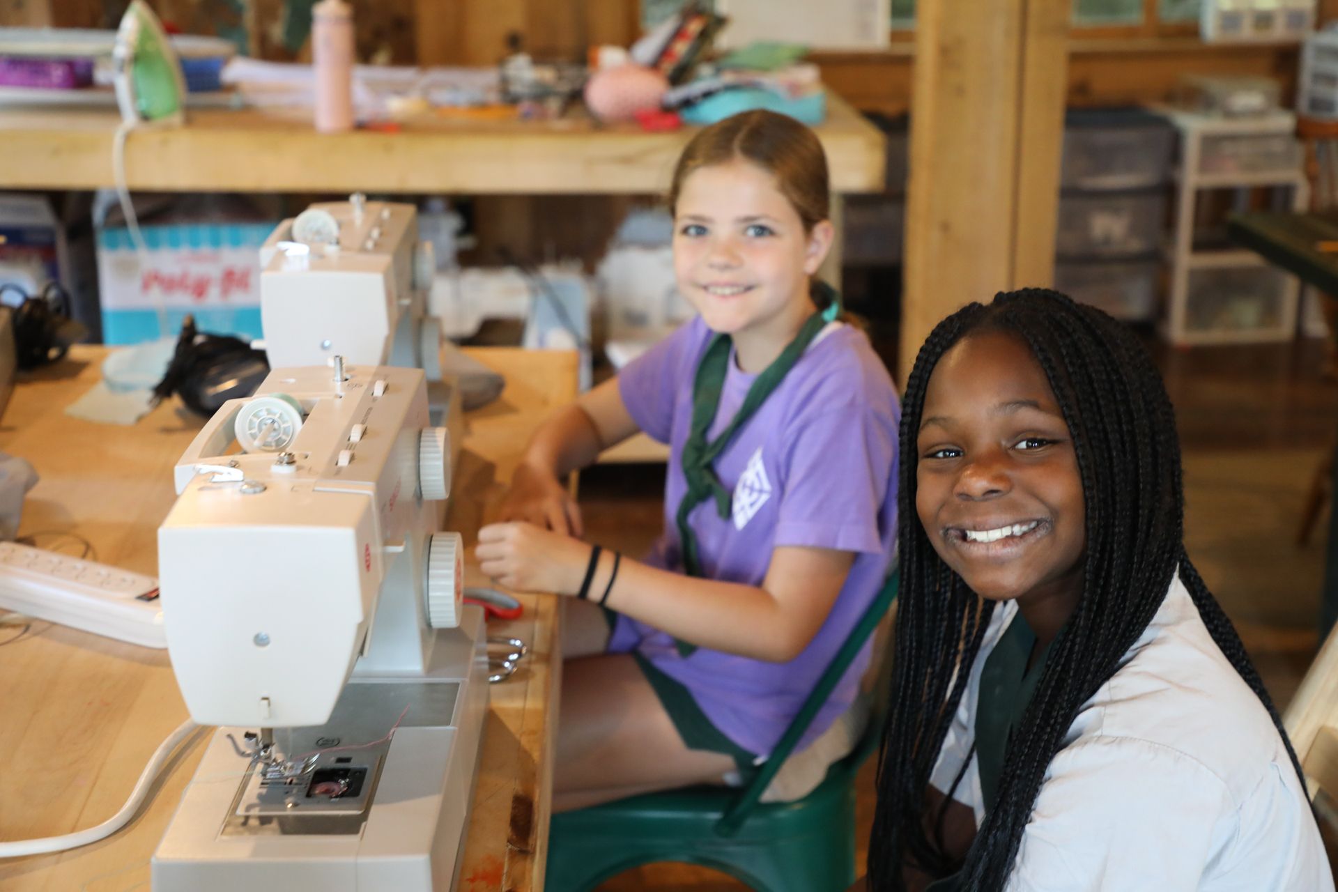 Two people smile while using sewing machines at a craft table in a bright workshop.