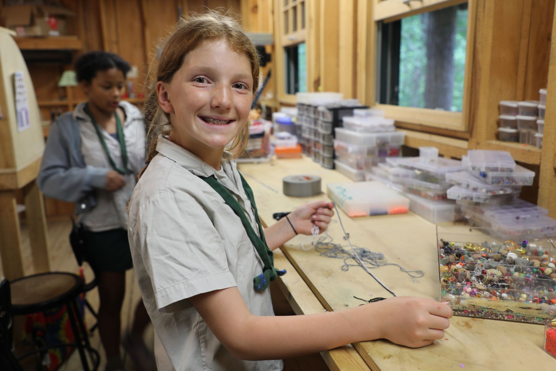 A person smiling while threading beads on a string at a wooden craft table, with another person in the background.