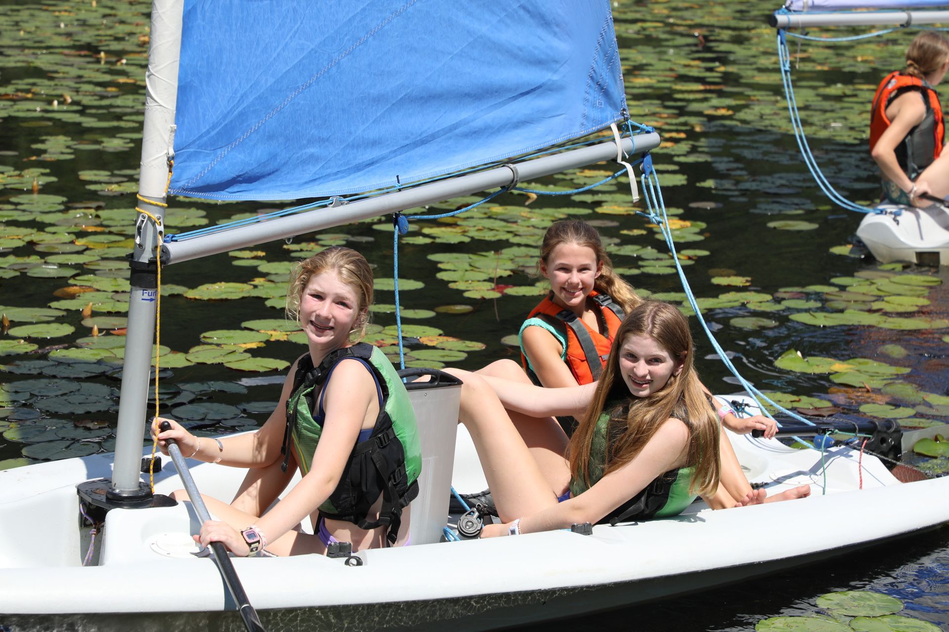 Three people in life jackets sail a small white boat with a blue sail on a water-lily-covered lake.