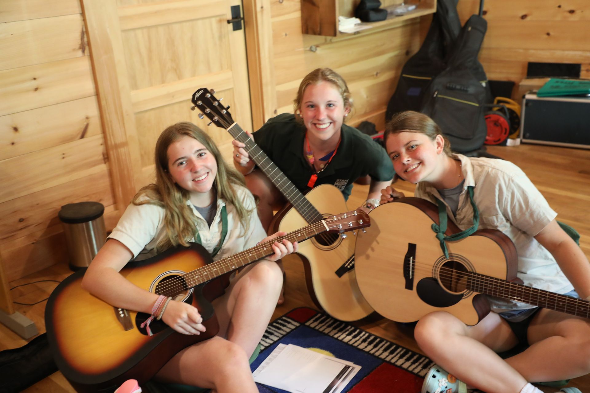 Three smiling people sitting on a wooden floor in a room, each holding an acoustic guitar.