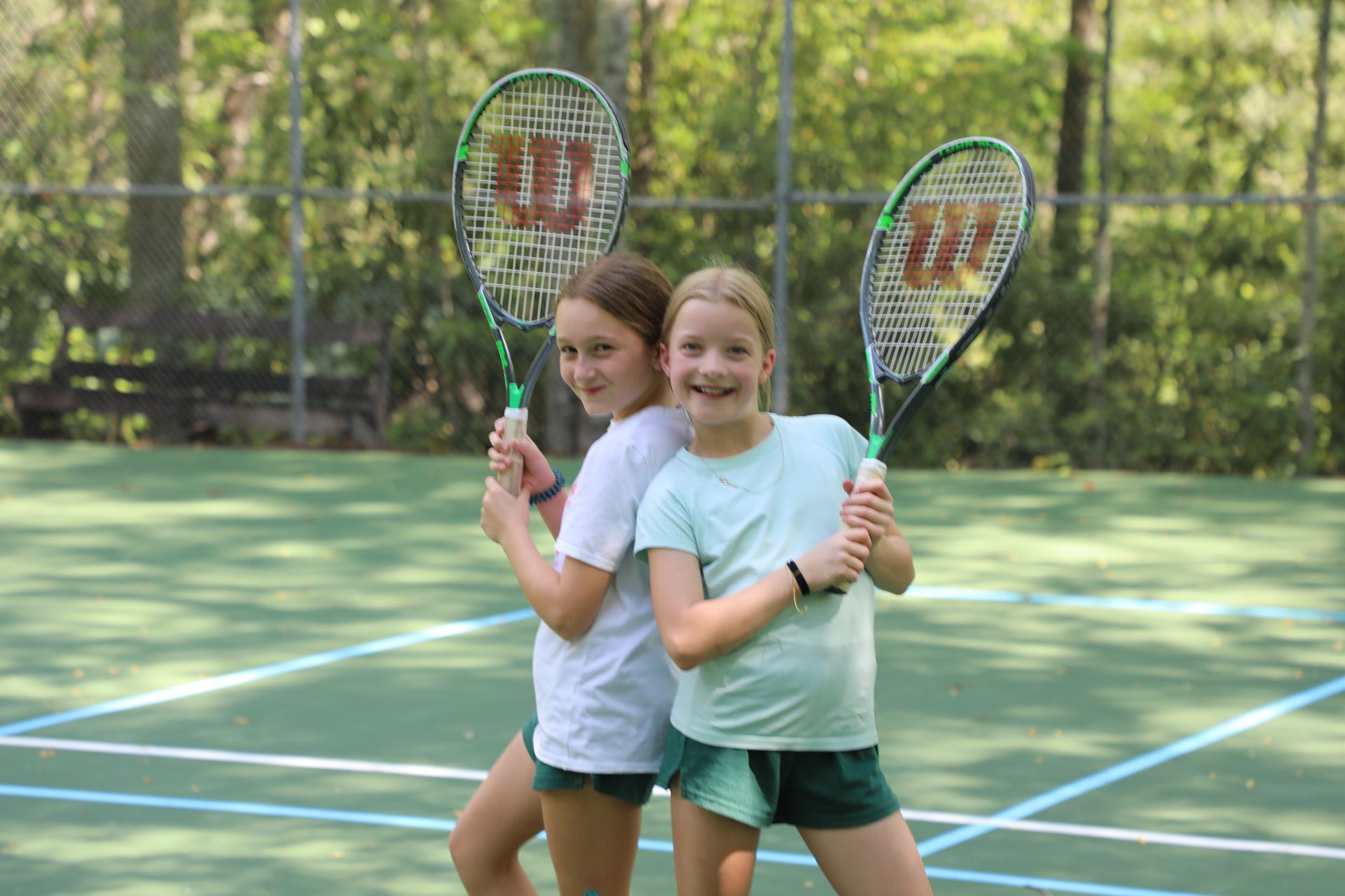 Two people stand back-to-back on a tennis court, holding Wilson rackets and smiling at the camera.