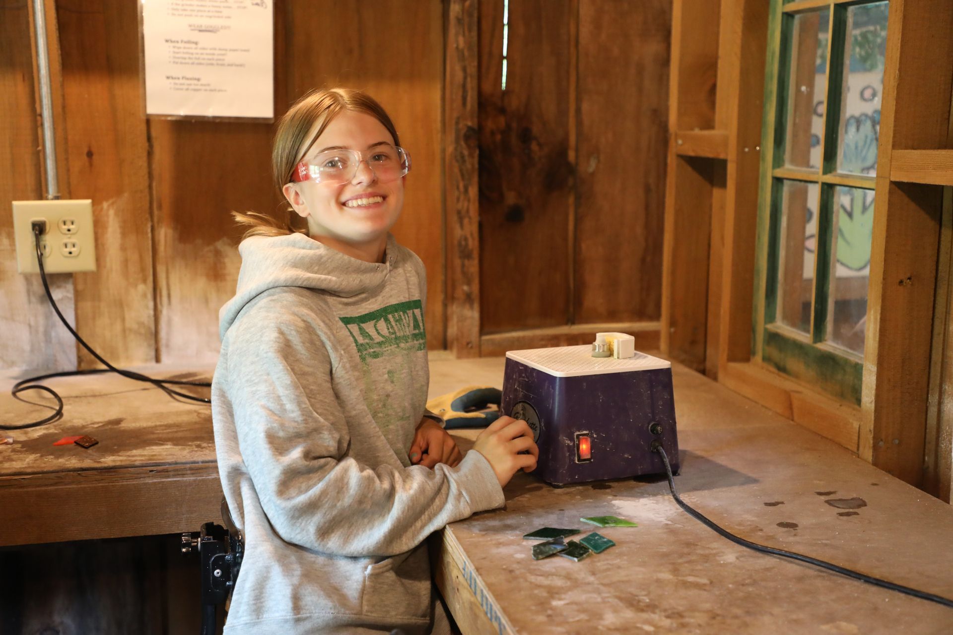 A person in safety glasses works at a grinder machine in a workshop with wooden walls and windows.