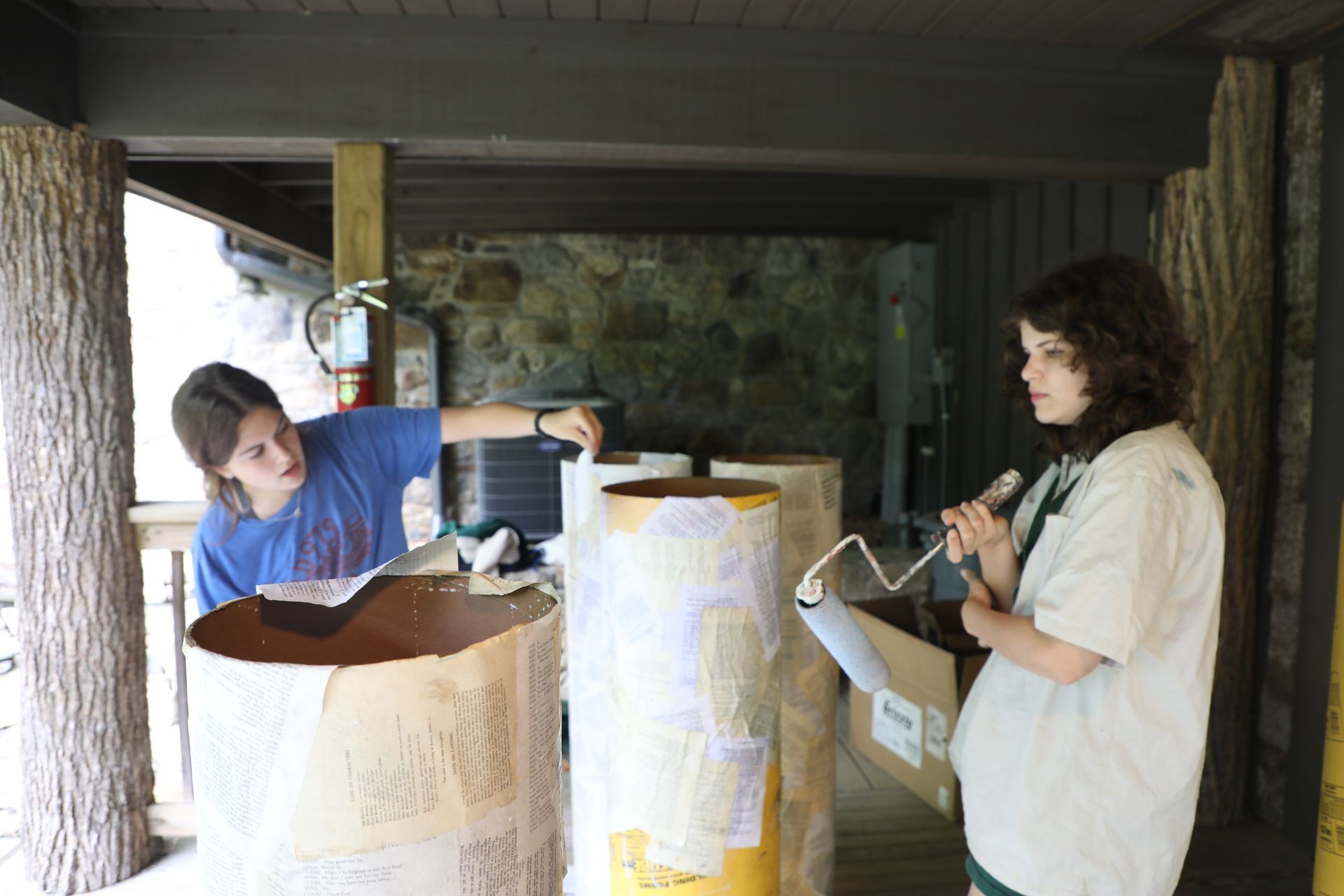 Two people work on a craft project, applying paper to cardboard columns under a stone-walled outdoor shelter.