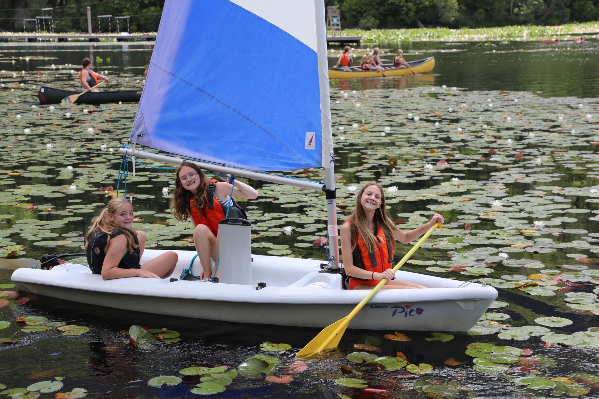 Three people in life jackets sail a small white boat with a blue and white sail through a lily-covered lake.