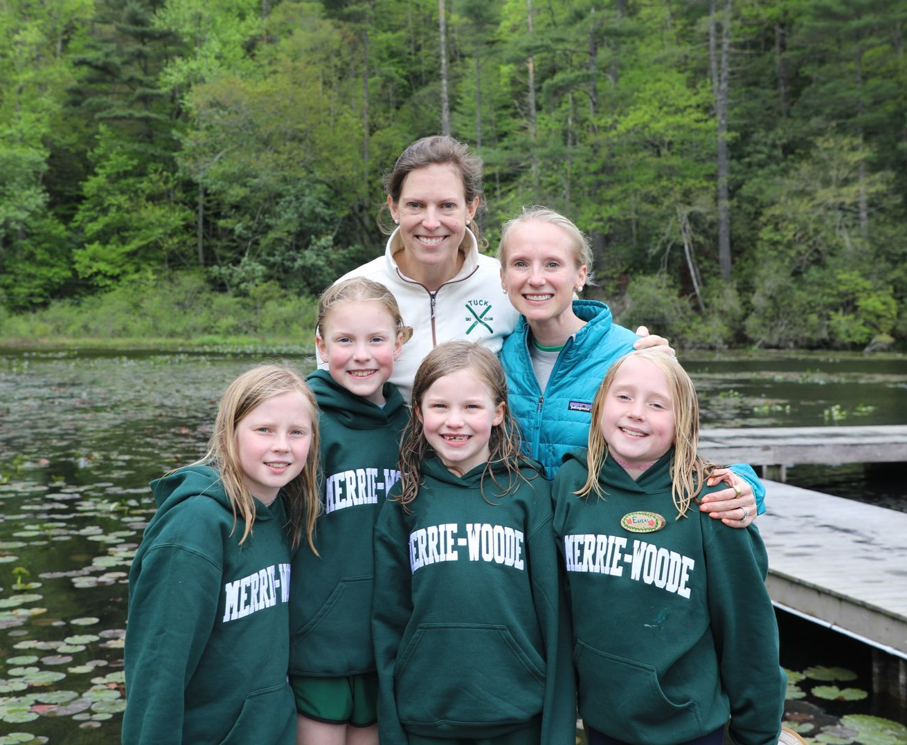 A group of six people wearing matching dark green Merrie-Woode hoodies smile in front of a lake and forest.
