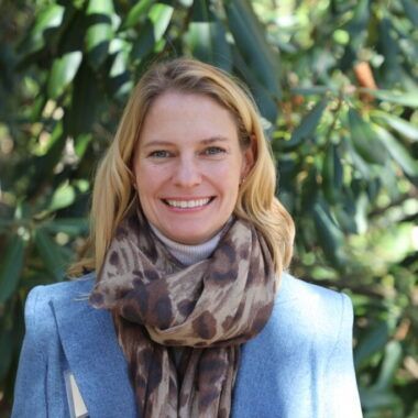 A person smiling, wearing a blue blazer, white turtleneck, and leopard-print scarf, standing in front of green foliage.