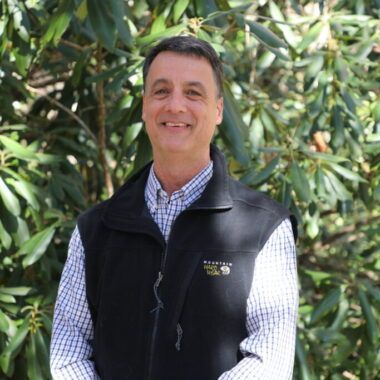 A smiling man wearing a plaid shirt and dark fleece vest stands in front of a green leafy backdrop.