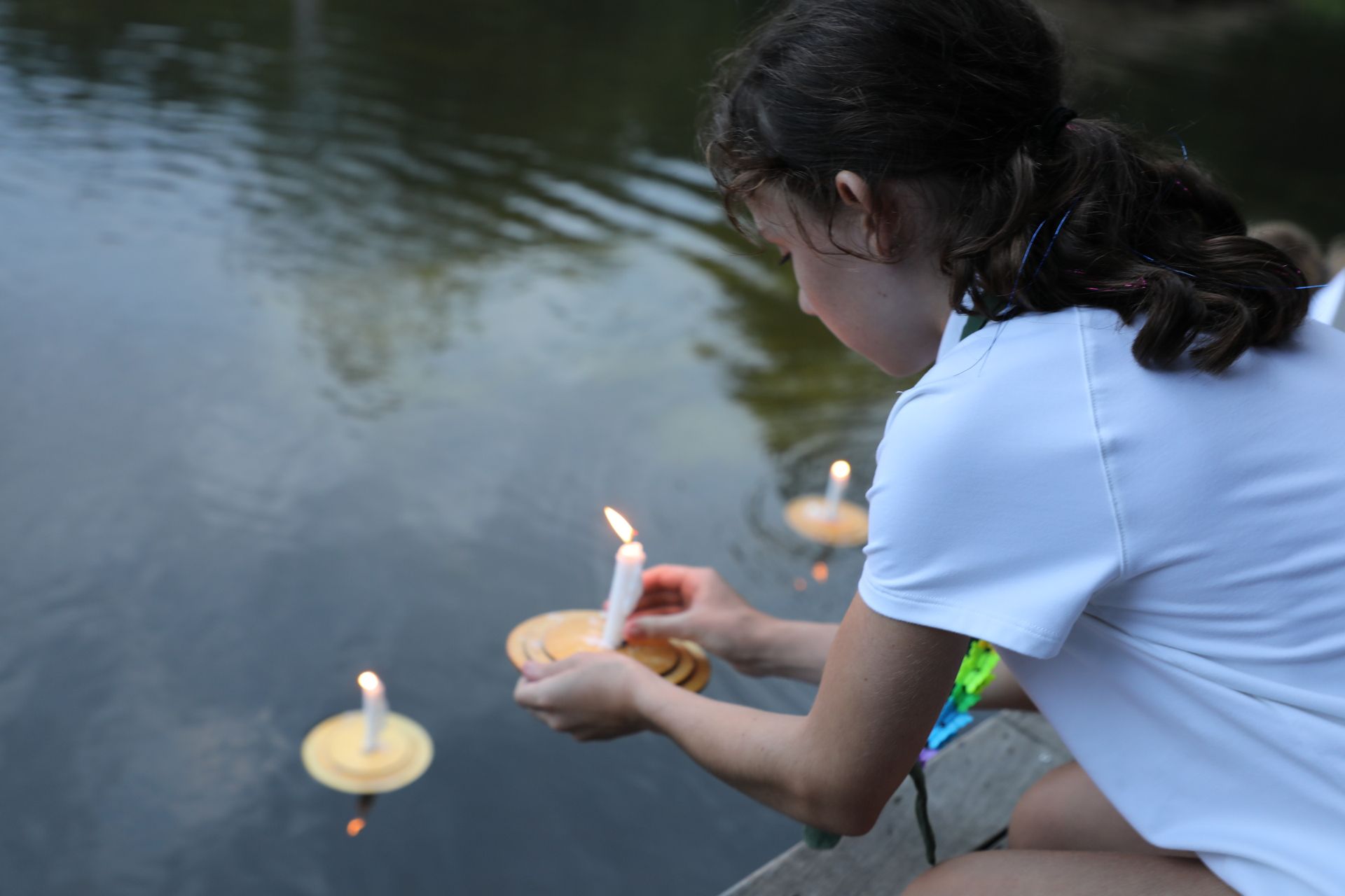 A person in a white shirt kneeling by a lake, placing a lit candle on a floating disk into the water.