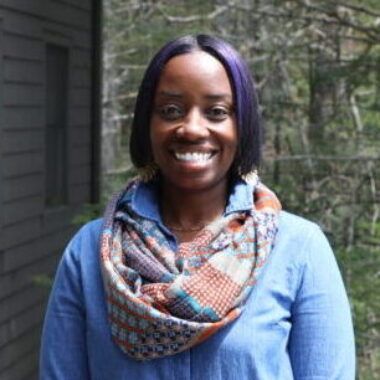 A smiling person wearing a blue shirt and a colorful patterned infinity scarf, standing outdoors in front of trees.