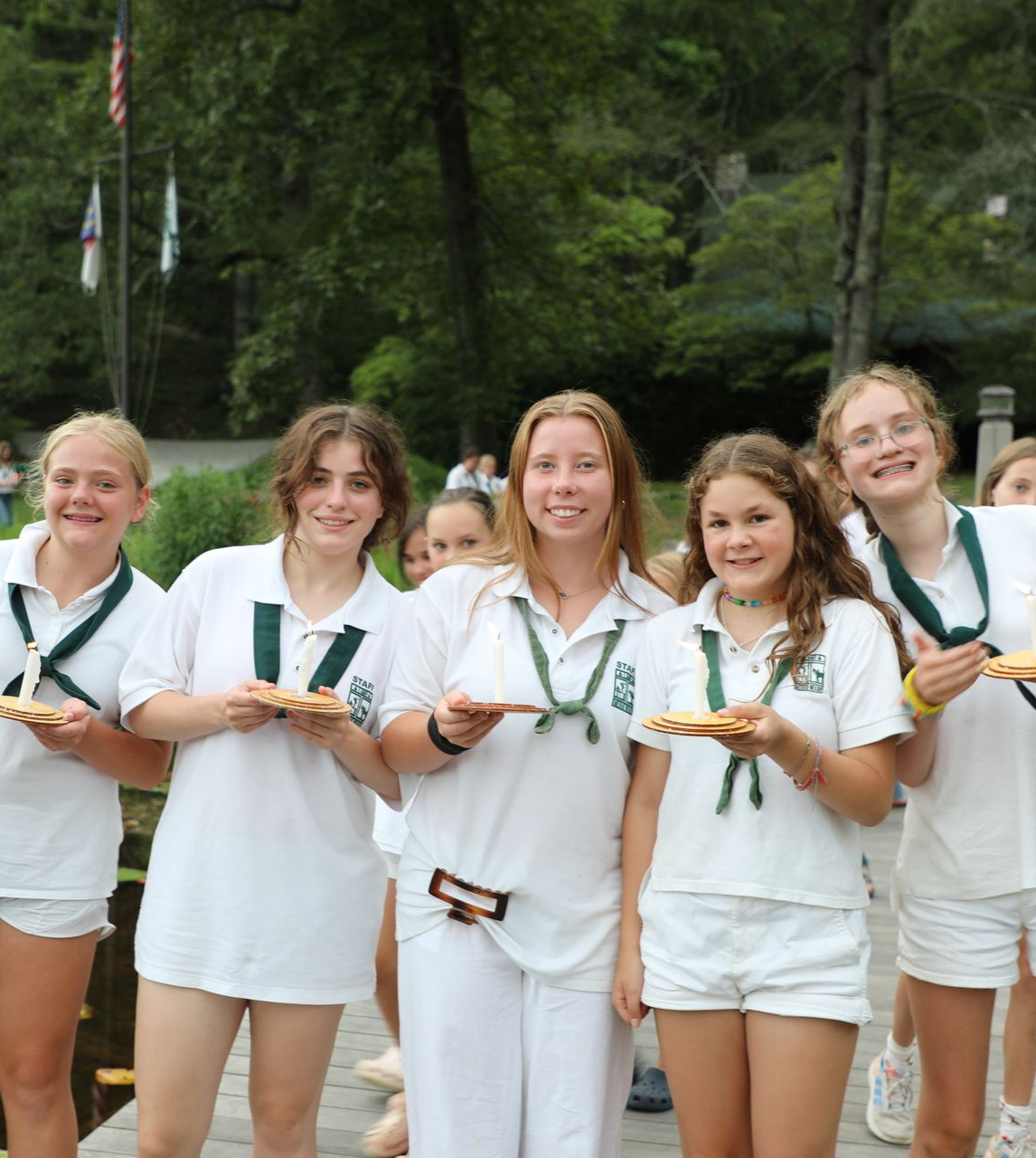 A group of people wearing white polo shirts and green scarves stand together outdoors while eating slices of pizza.