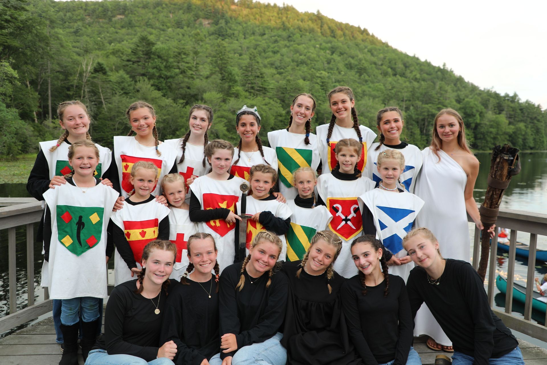 A large group of people in medieval-style tunics with crests, standing on a wooden dock by a lake and wooded hill.