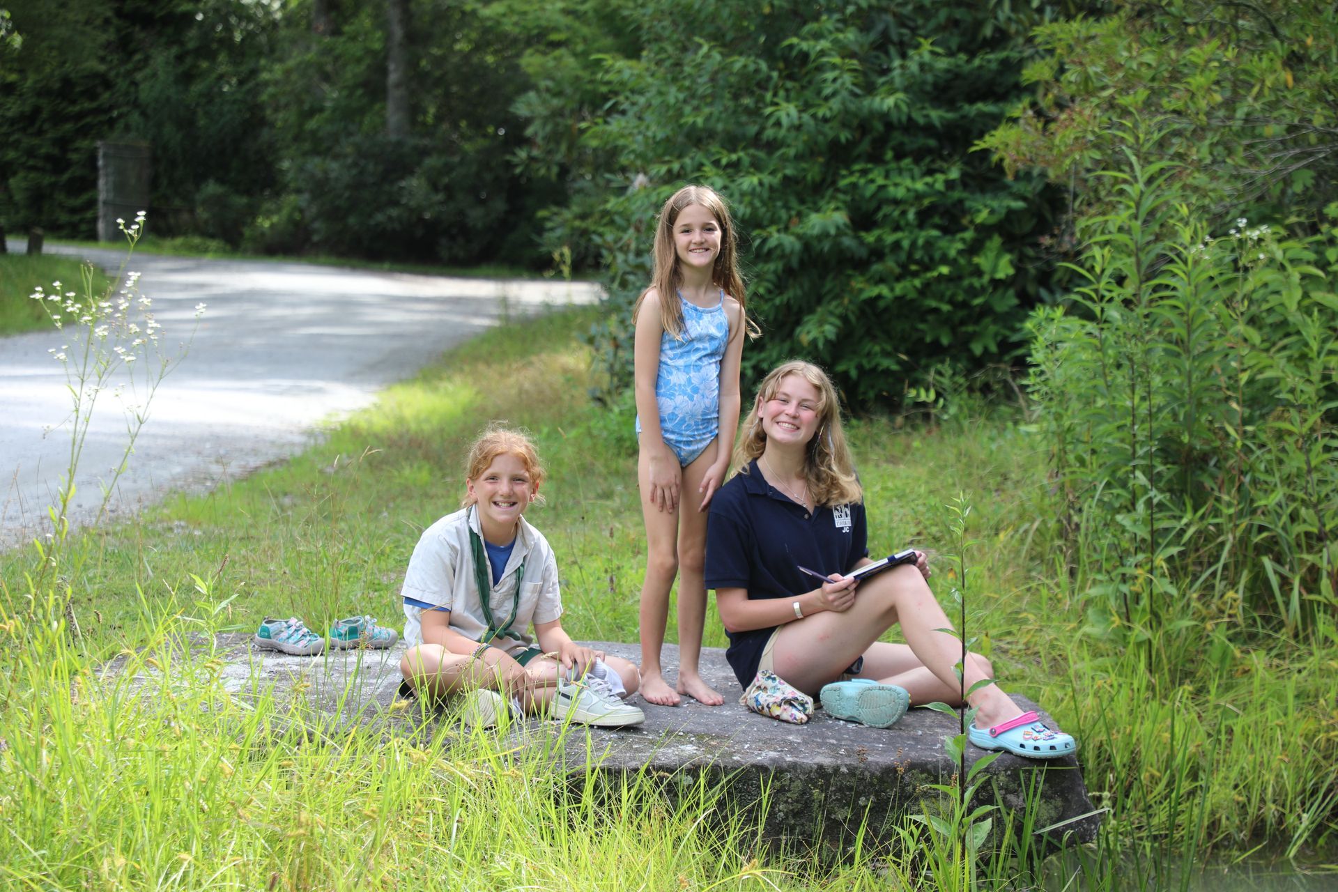 Three children pose together on a concrete ledge by a grassy path and water, smiling at the camera.