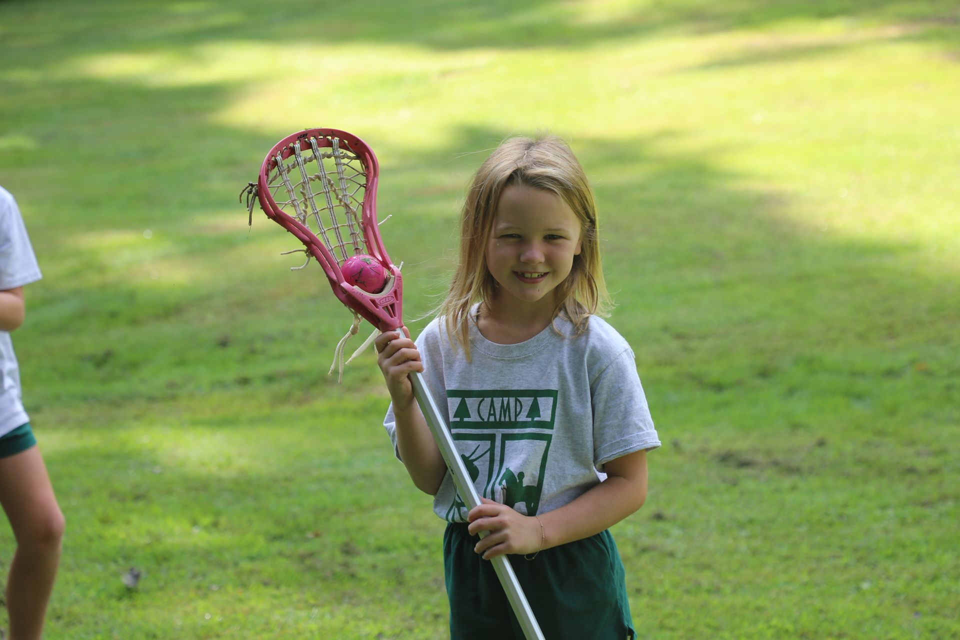 A smiling child stands on a grassy field holding a red lacrosse stick with a pink ball in the mesh pocket.