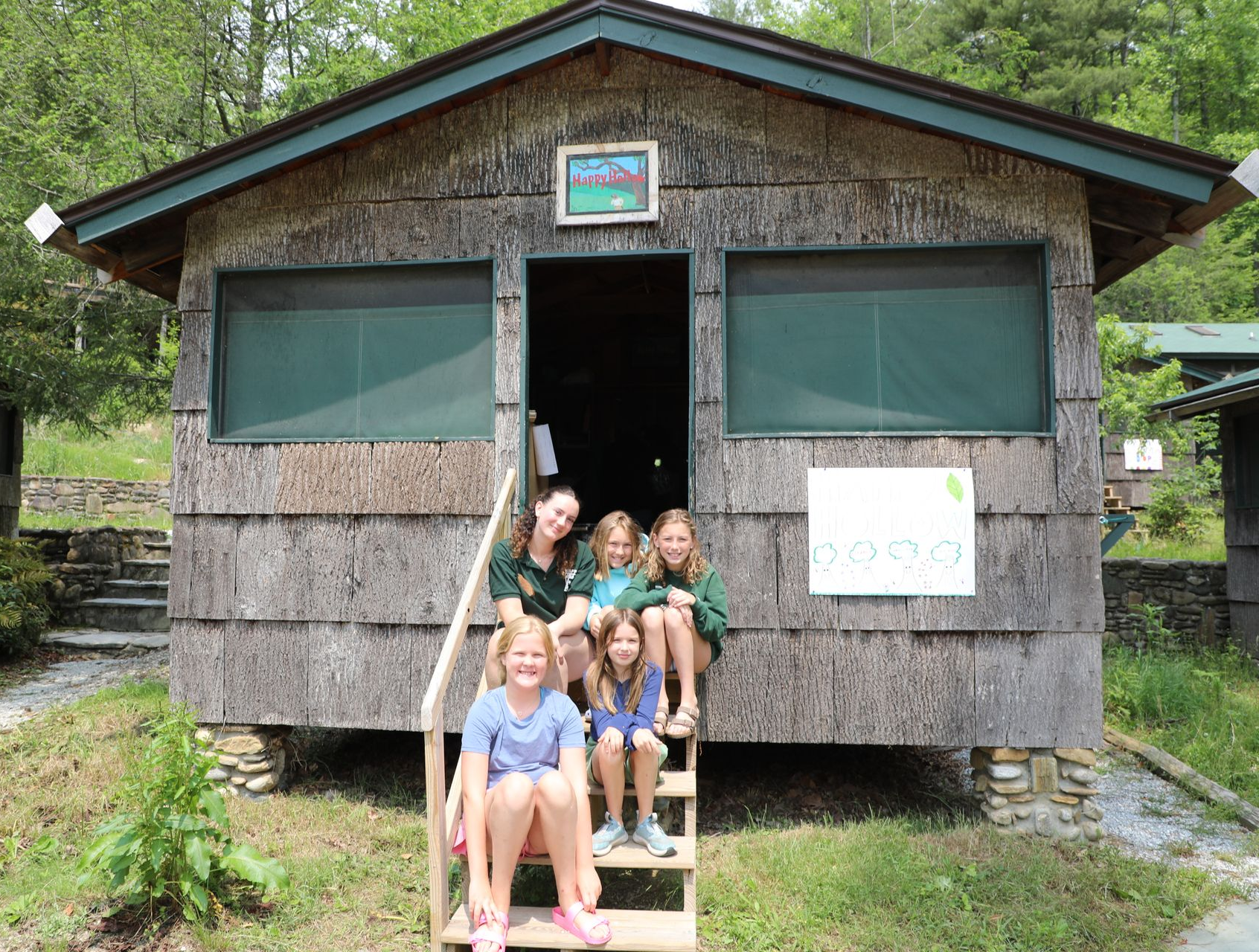 Five people pose on the stairs of a rustic wooden cabin surrounded by trees.