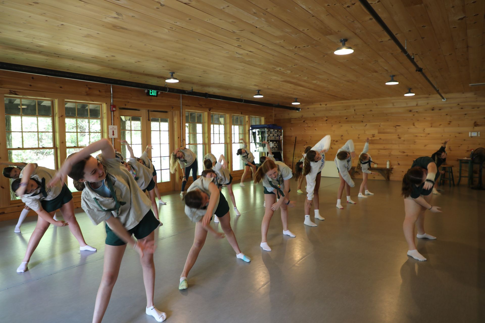 A group of people wearing matching uniforms practicing rhythmic stretching exercises in a wood-paneled dance studio.