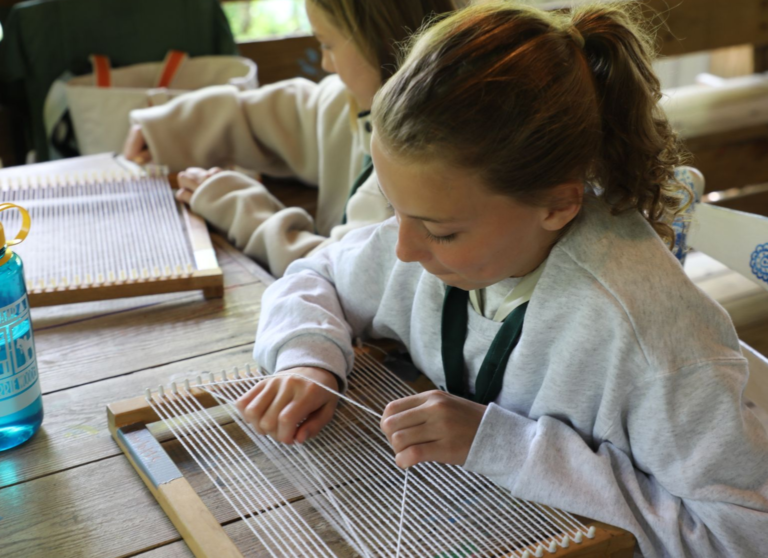 Two people focus intently on weaving with white yarn on wooden looms at a wooden table.