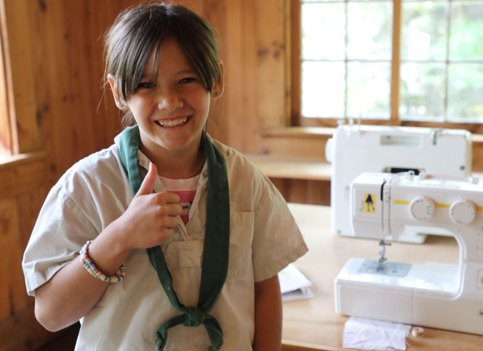 A smiling child in a scout uniform giving a thumbs up in a wooden room with a sewing machine on a table.