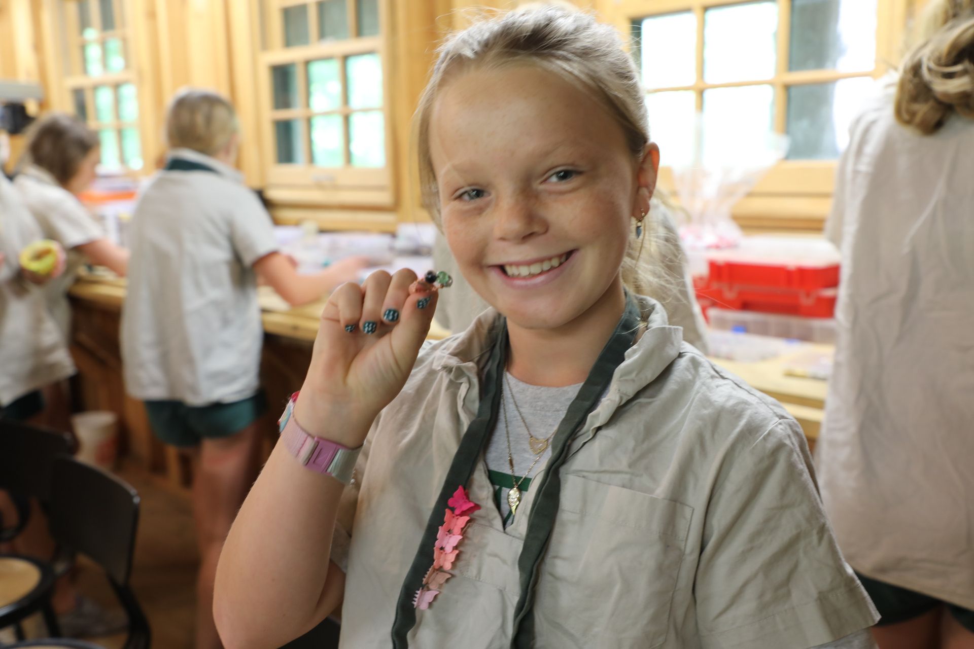 A smiling person indoors holds up a small, dark object while others work at a wooden table in the background.