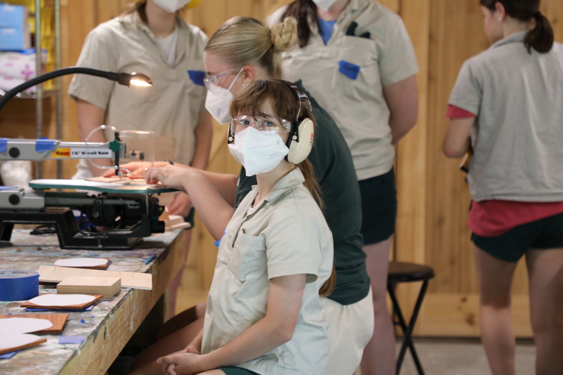 People wearing masks and safety gear use a scroll saw at a workbench in a woodshop.