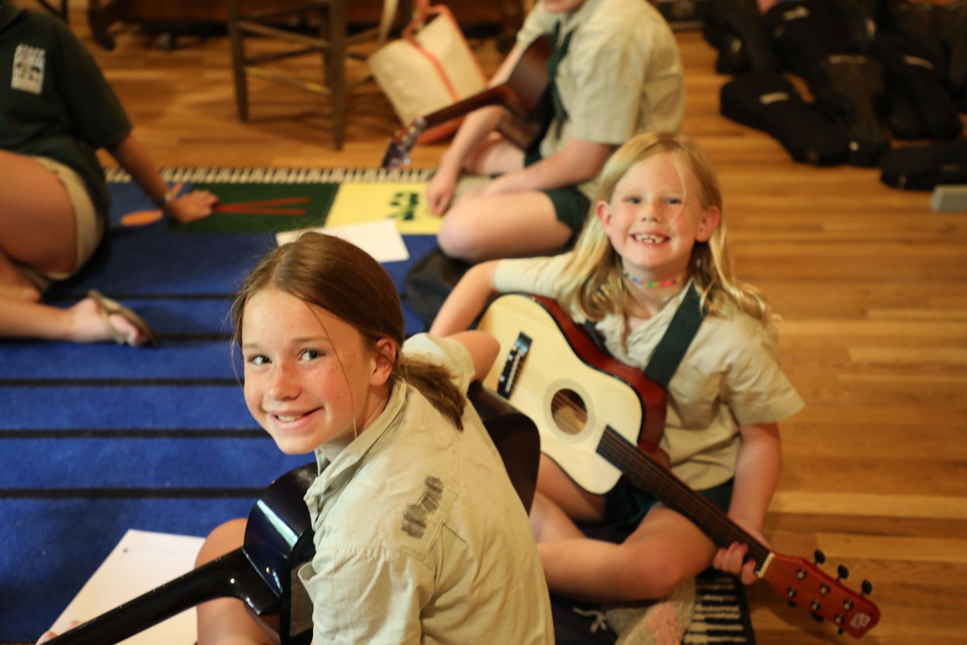 Two children in uniform sitting on a blue rug, smiling while holding acoustic guitars.